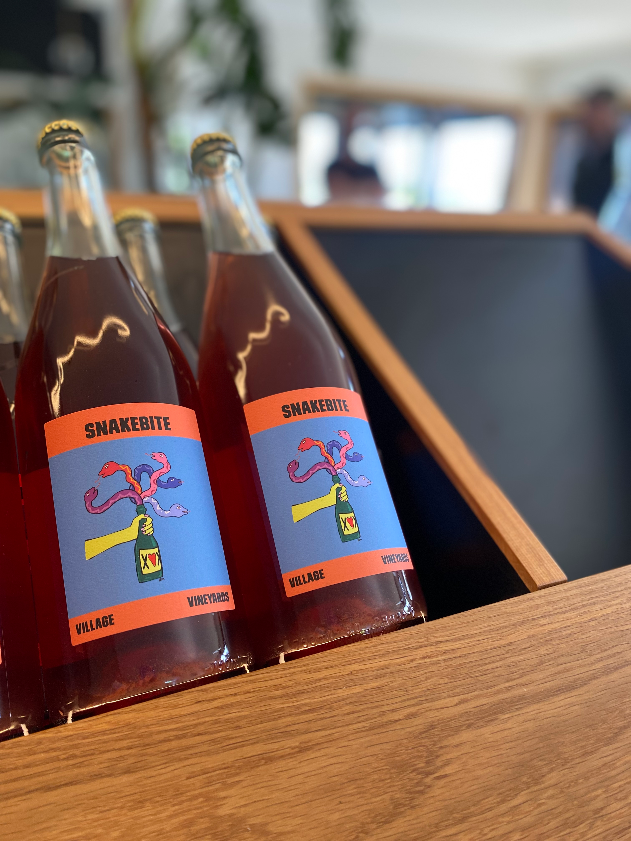 Four bottles of Snakebite beverage on a wooden surface at a bar or restaurant, with a blurred background of people and furniture. Custom wine bottle display unit solutions