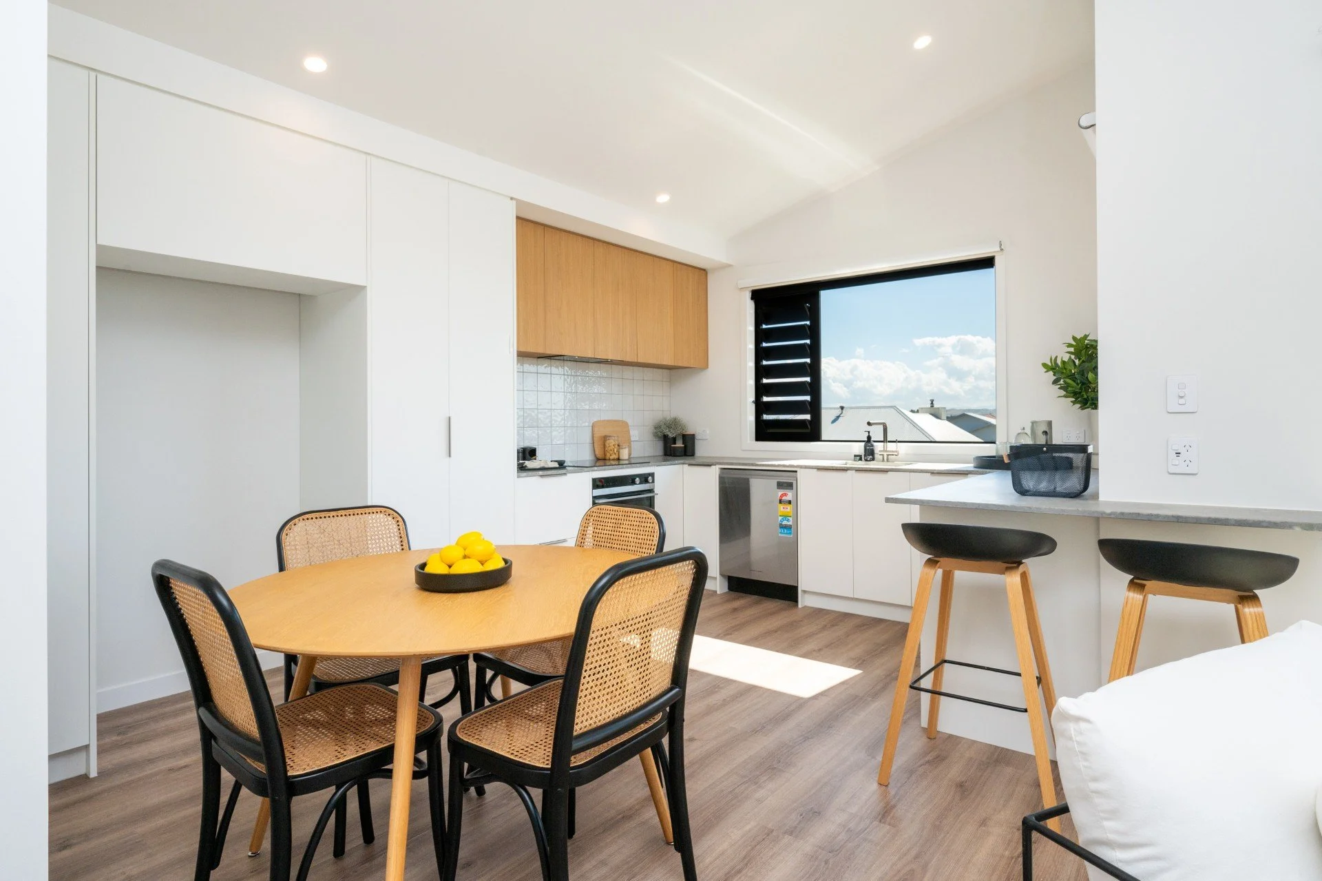 Modern kitchen and dining area with wooden table, black and brown chairs, white cabinets, and a window showing a blue sky with clouds.
