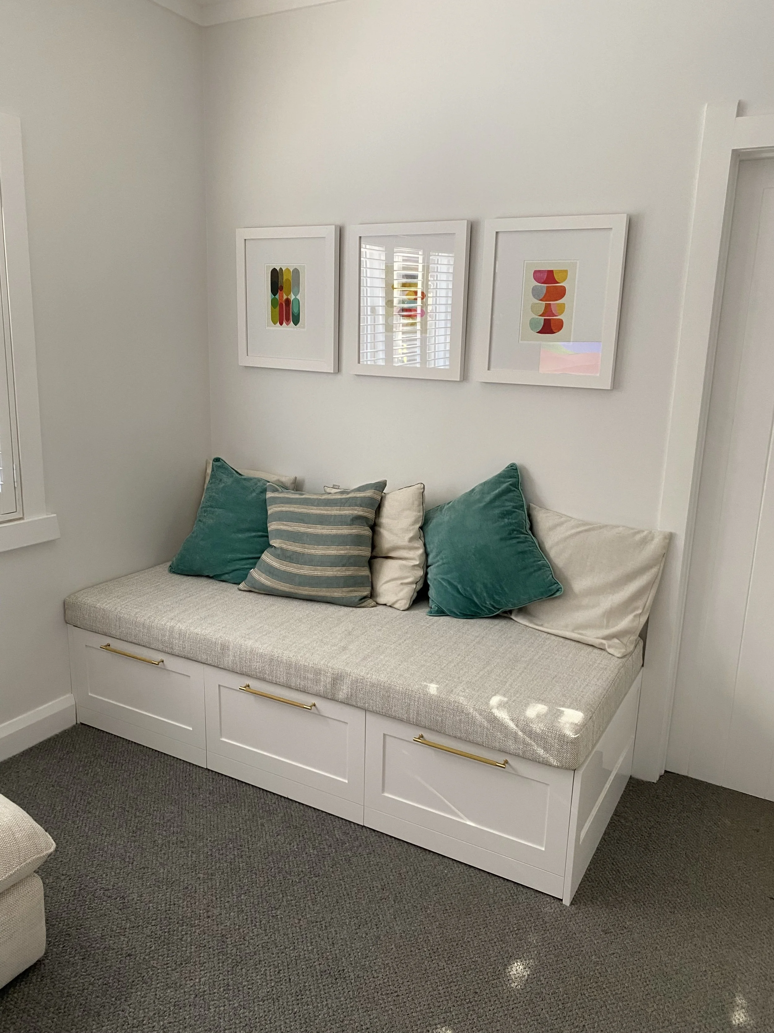 A cozy corner with a built-in white storage bench with drawers underneath, topped with a cushioned seat and five throw pillows in shades of teal, white, and striped pattern. Above, three framed abstract art prints hang on a white wall, with a window featuring white plantation shutters reflected in the middle picture.