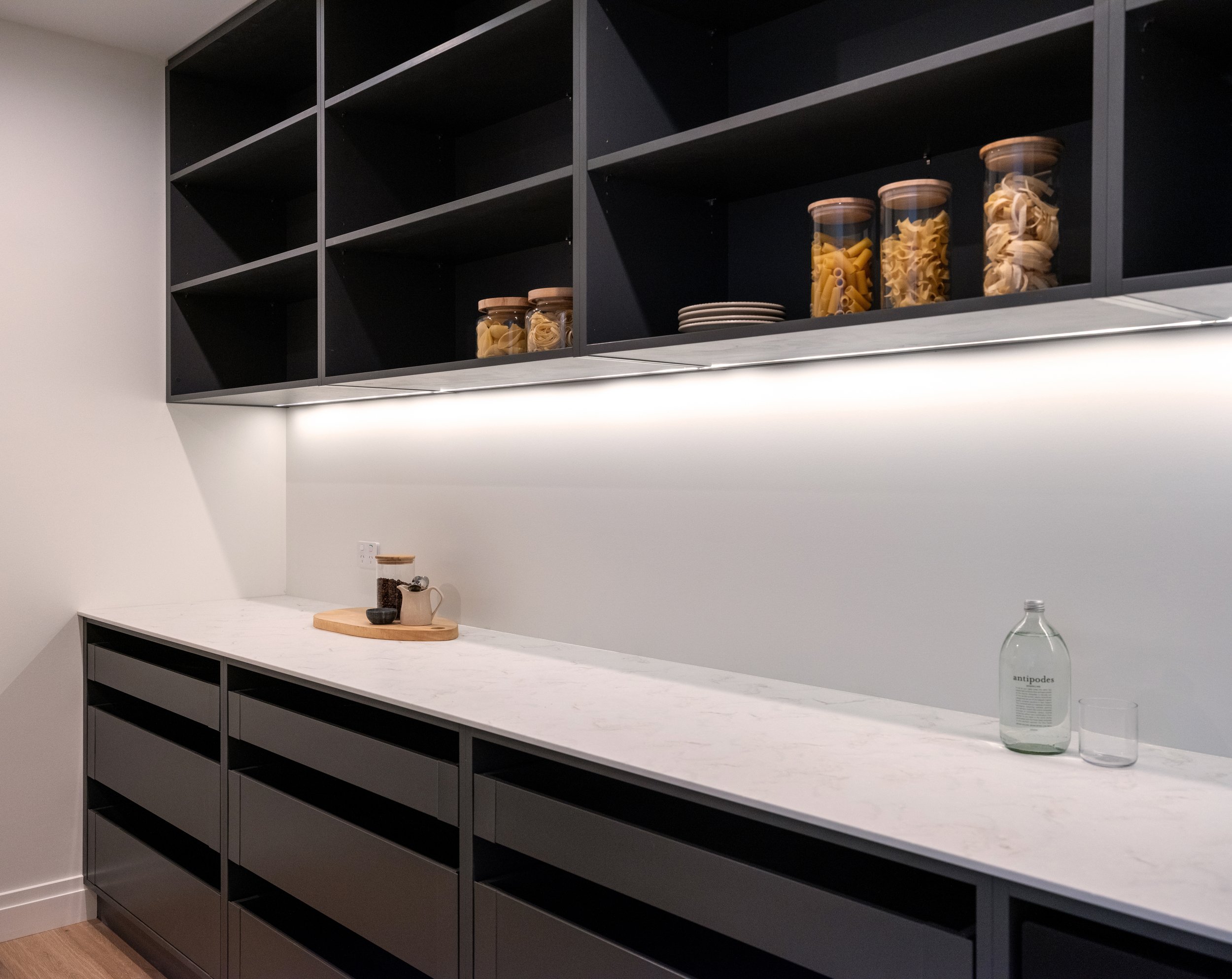 Kitchen with black open shelves and a white marble countertop, containing a small cutting board, jars, and a bottle of Antipodes water.