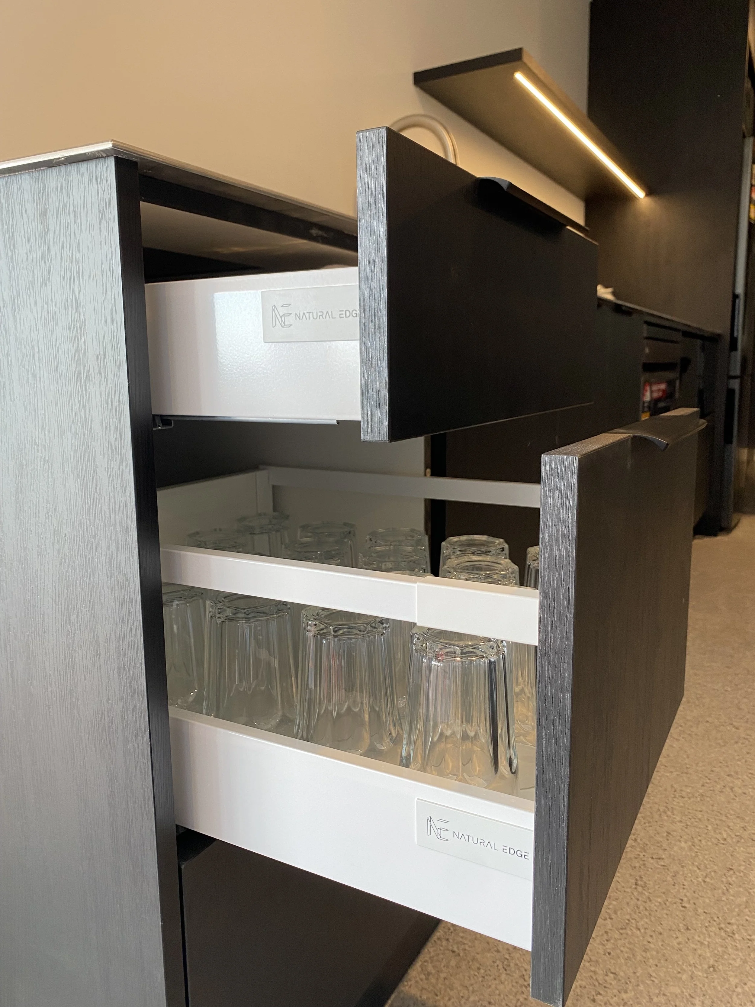 Open cabinet drawer containing overturned glasses on white tray with logo 'NATURAL EDGE' in a modern kitchen or bar area.