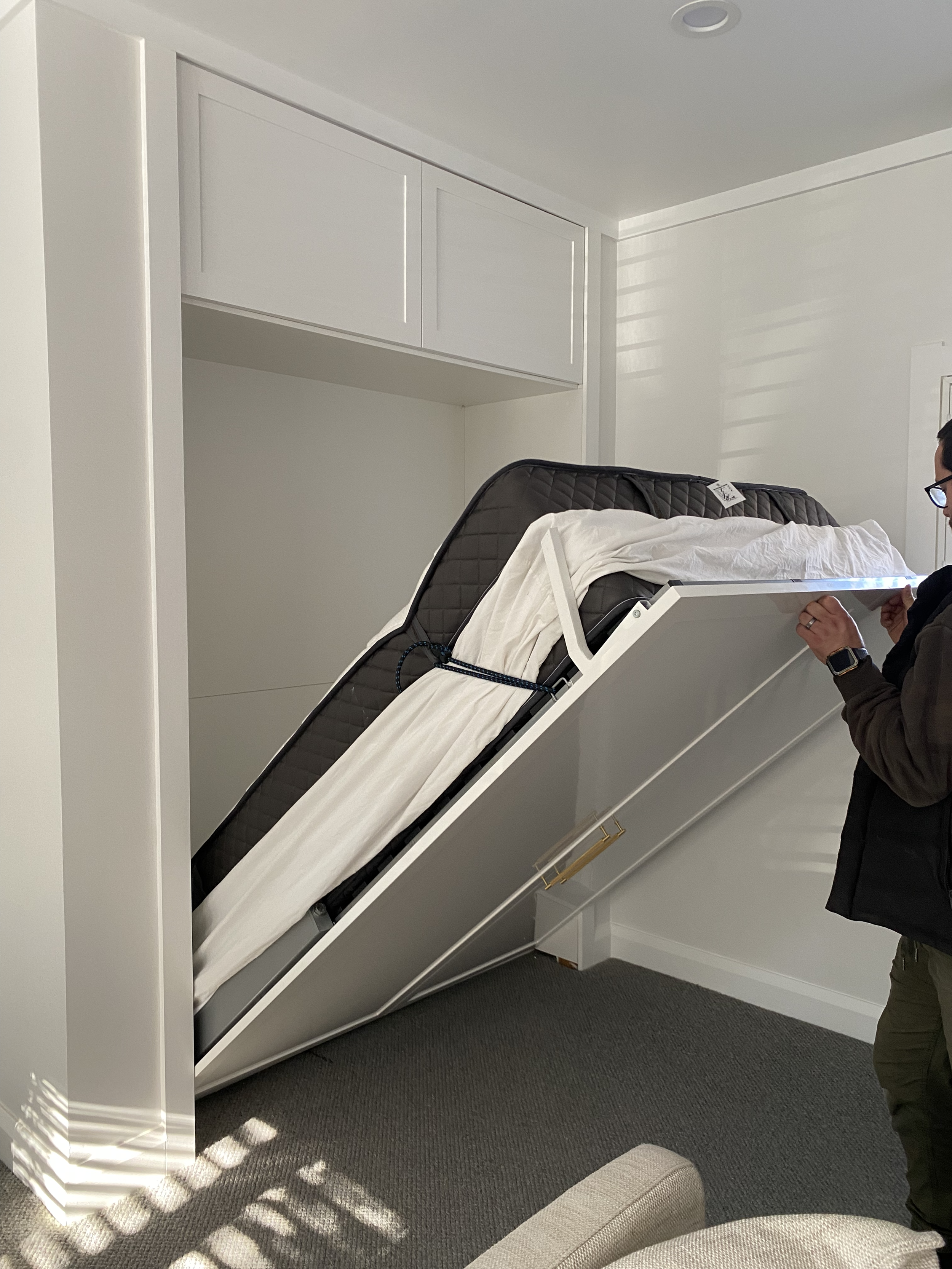 A person is standing next to a Murphy bed that is partially folded down, revealing a mattress with bedding on top of it. Pull-down bed for extra guests to stay