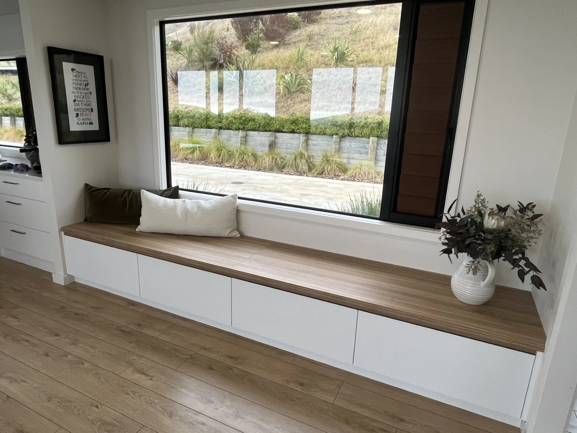 Interior of a room with a large window seat featuring a white and wood-tone cabinet beneath the window, decorated with a white pillow and a brown pillow, a white vase with dark foliage and flowers, and a framed art print on the wall. Outside the window, a landscaped yard with plants and a wooden retaining wall is visible.