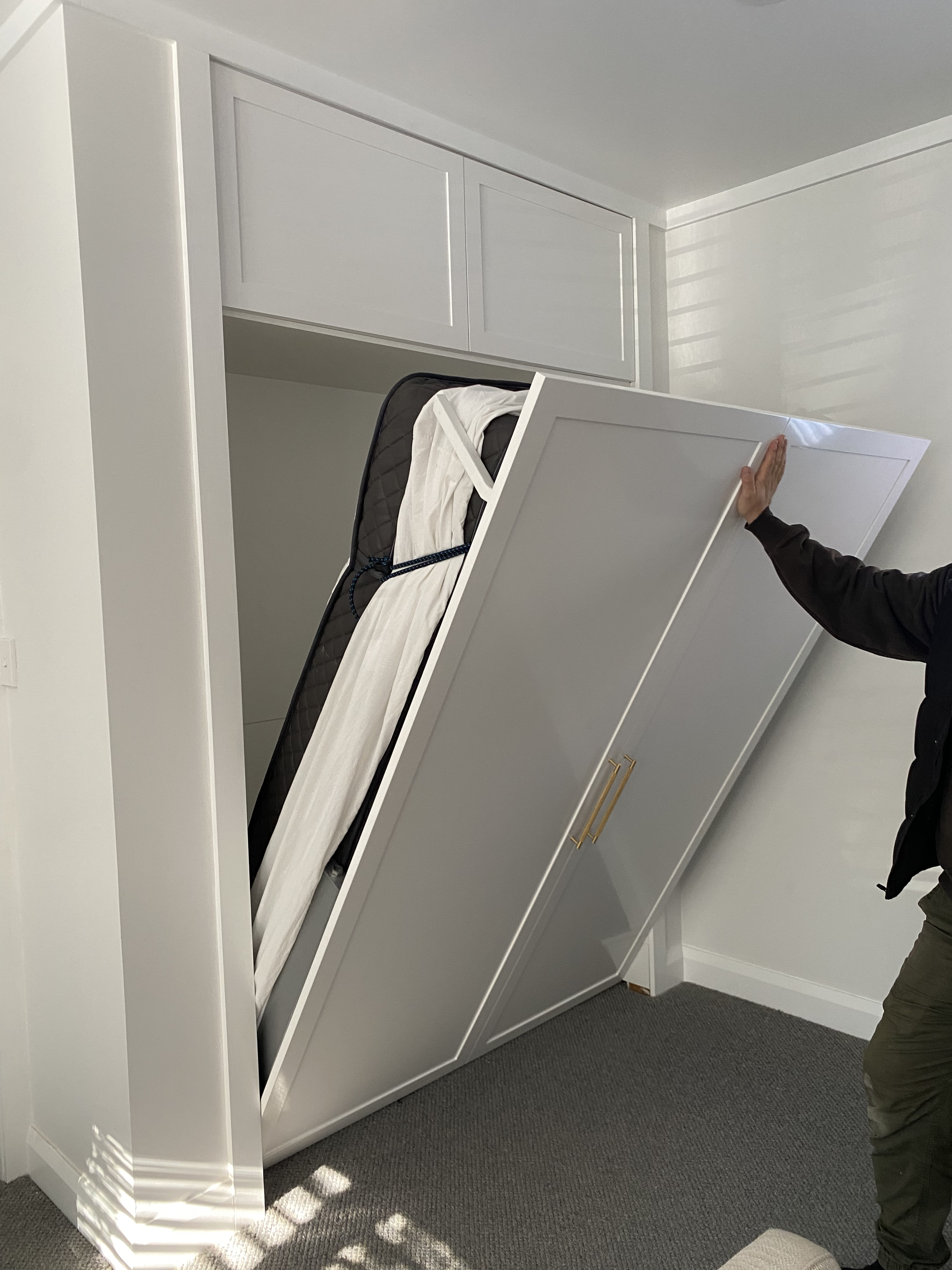 Person moving a white wardrobe to reveal a hidden bed with mattress inside a closet