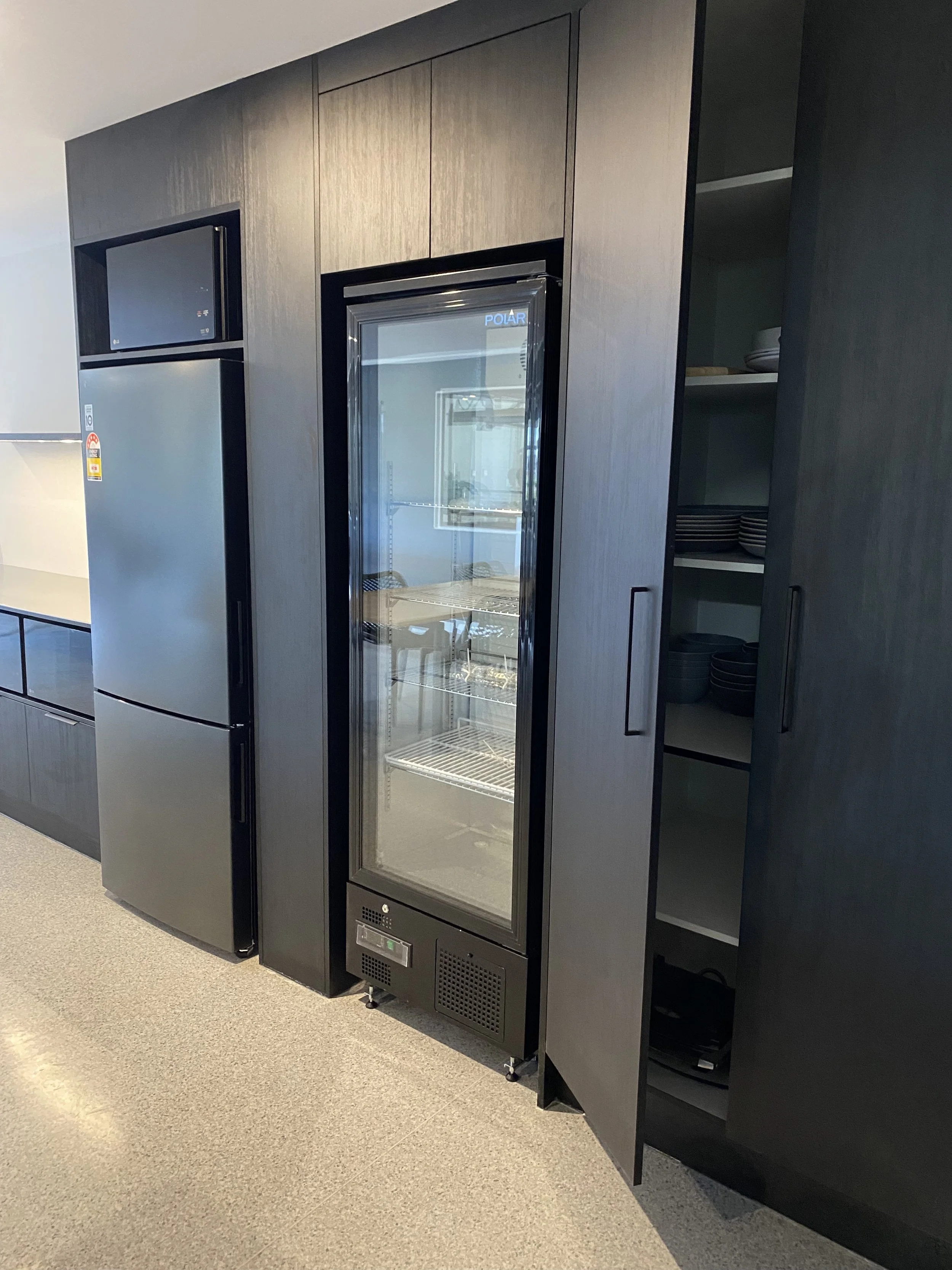 A black refrigerator with a glass door and a smaller black microwave above it, set into a dark wood cabinet with shelves and an open door revealing plates and bowls.