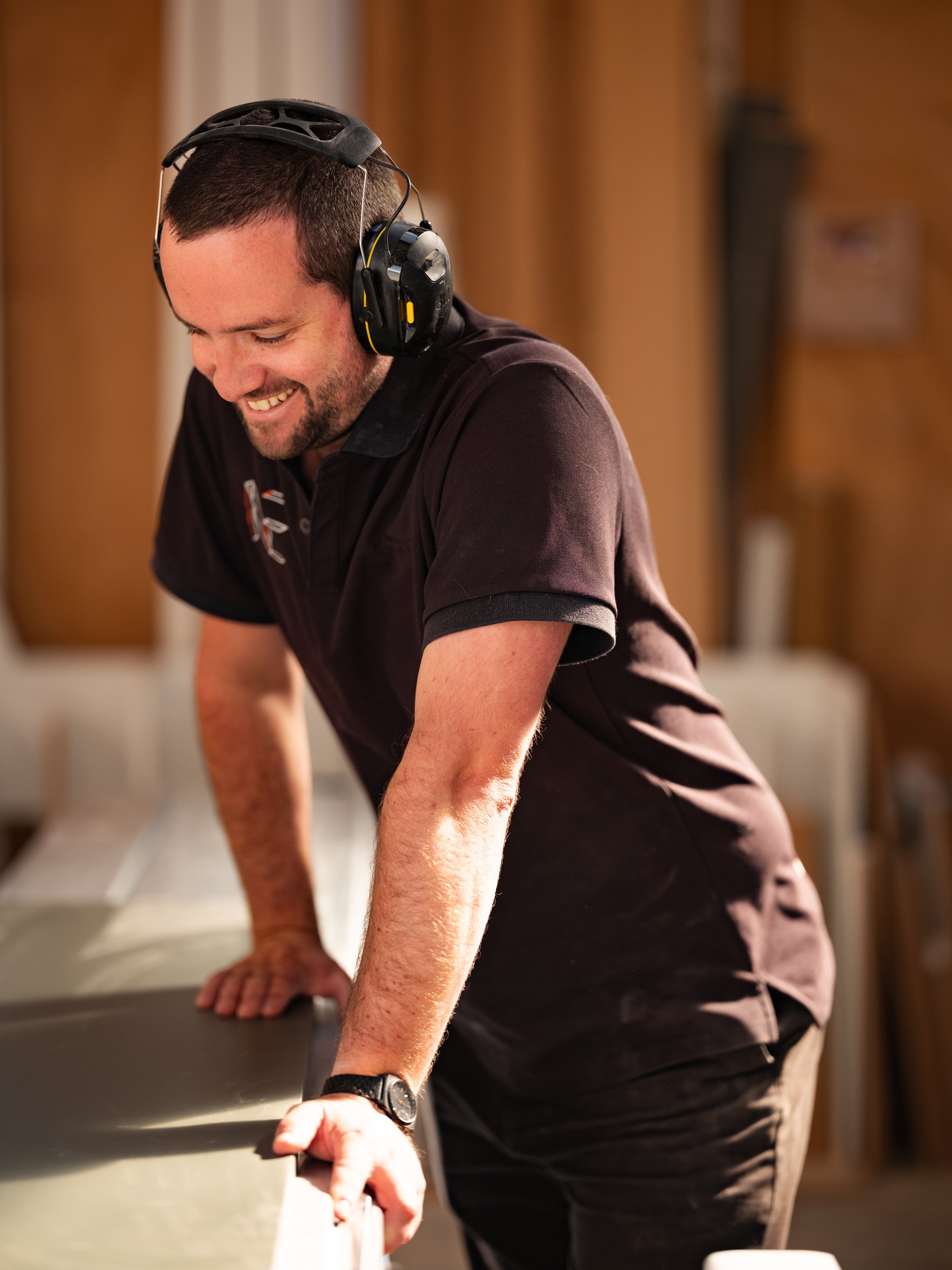 Man with short dark hair, beard, wearing black headphones, black watch, leaning on a table, smiling, in a room with brown curtains in the background.