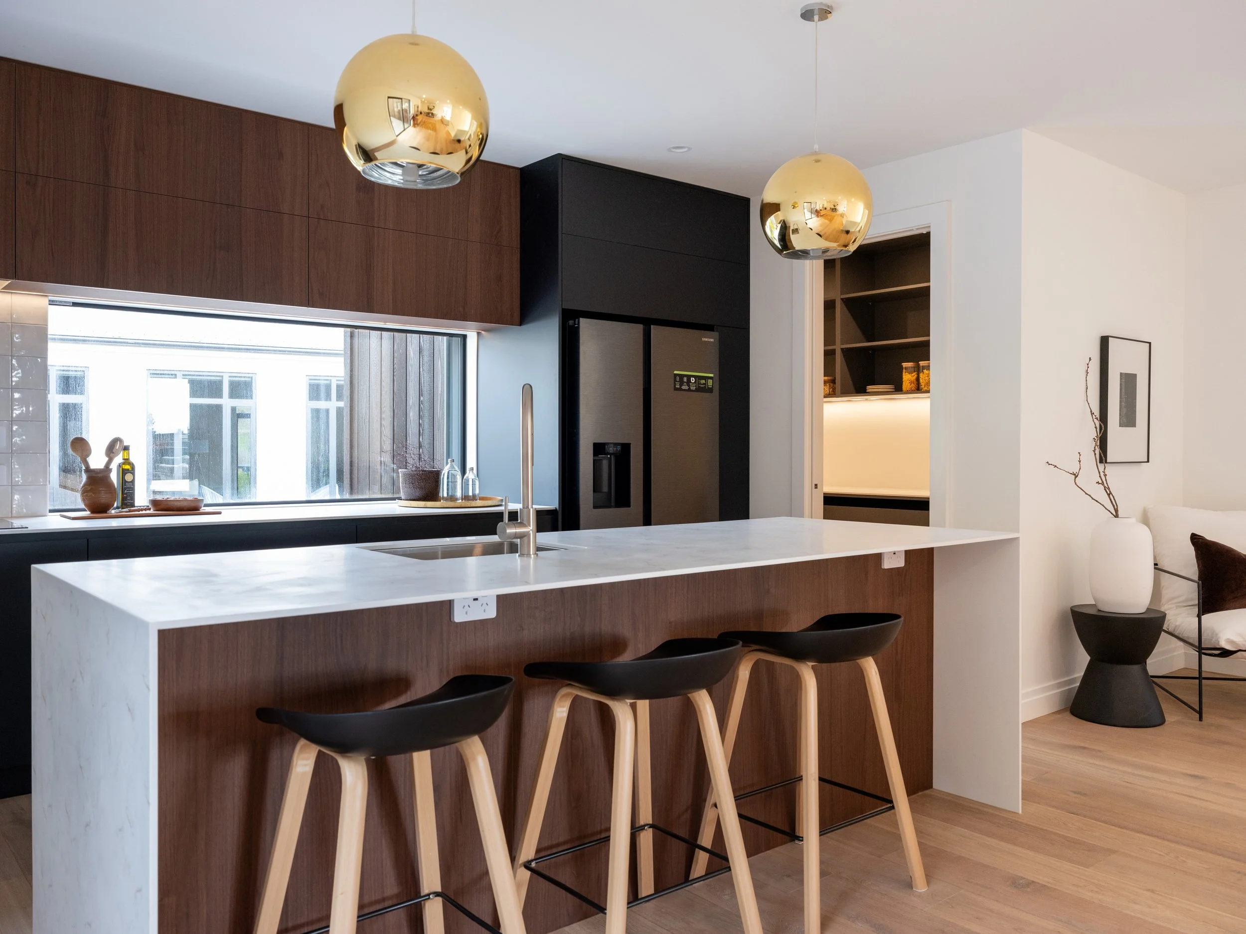 Modern kitchen with white marble island, black and wood cabinetry, gold pendant lights, and black bar stools with light wood legs.