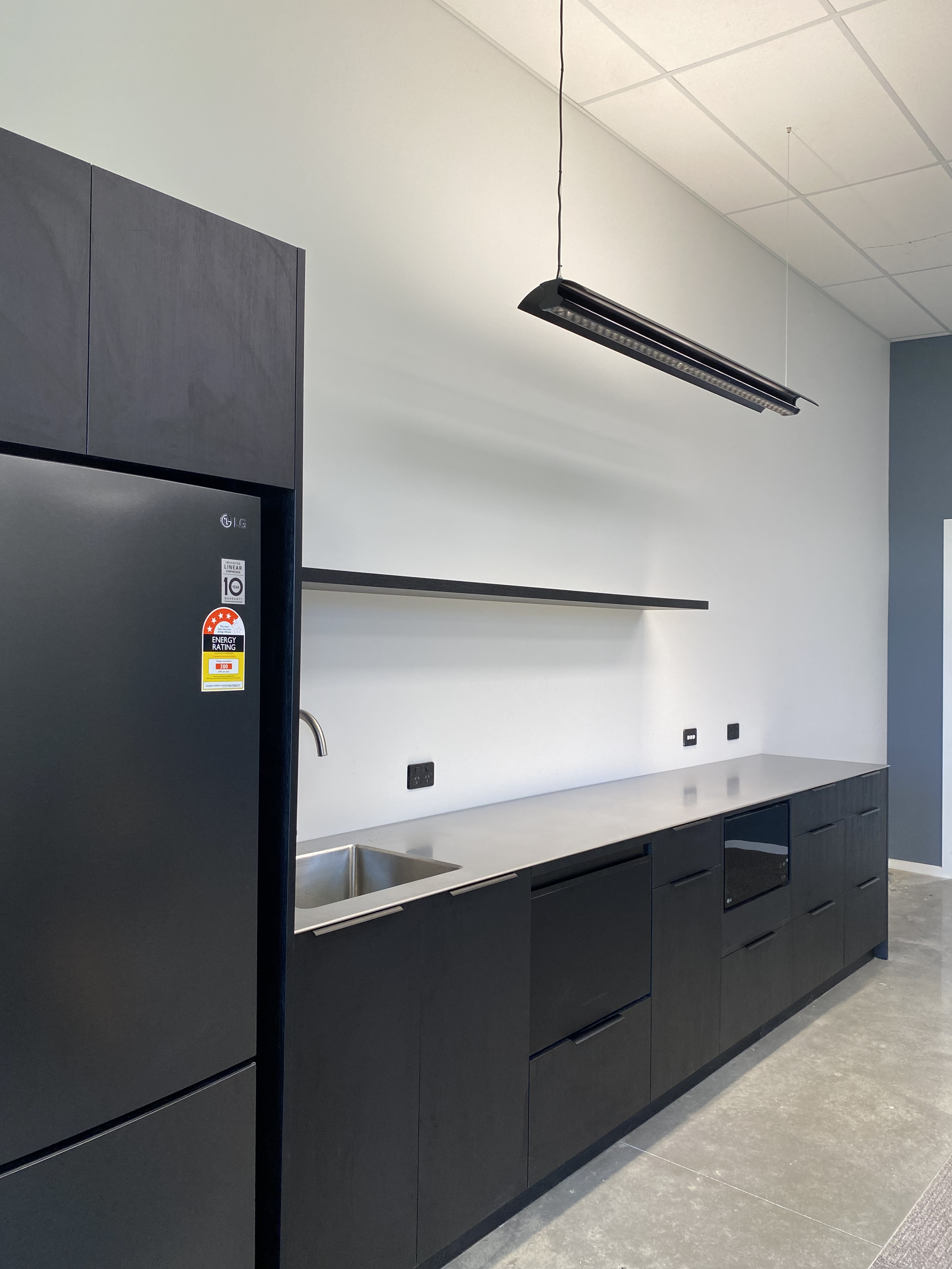 Modern staff kitchen with black cabinetry, a stainless steel sink, a black refrigerator, and a countertop, with black outlets on the white wall, and a suspended light fixture. Staff room joinery for seamless flow in the workplace break rooms.