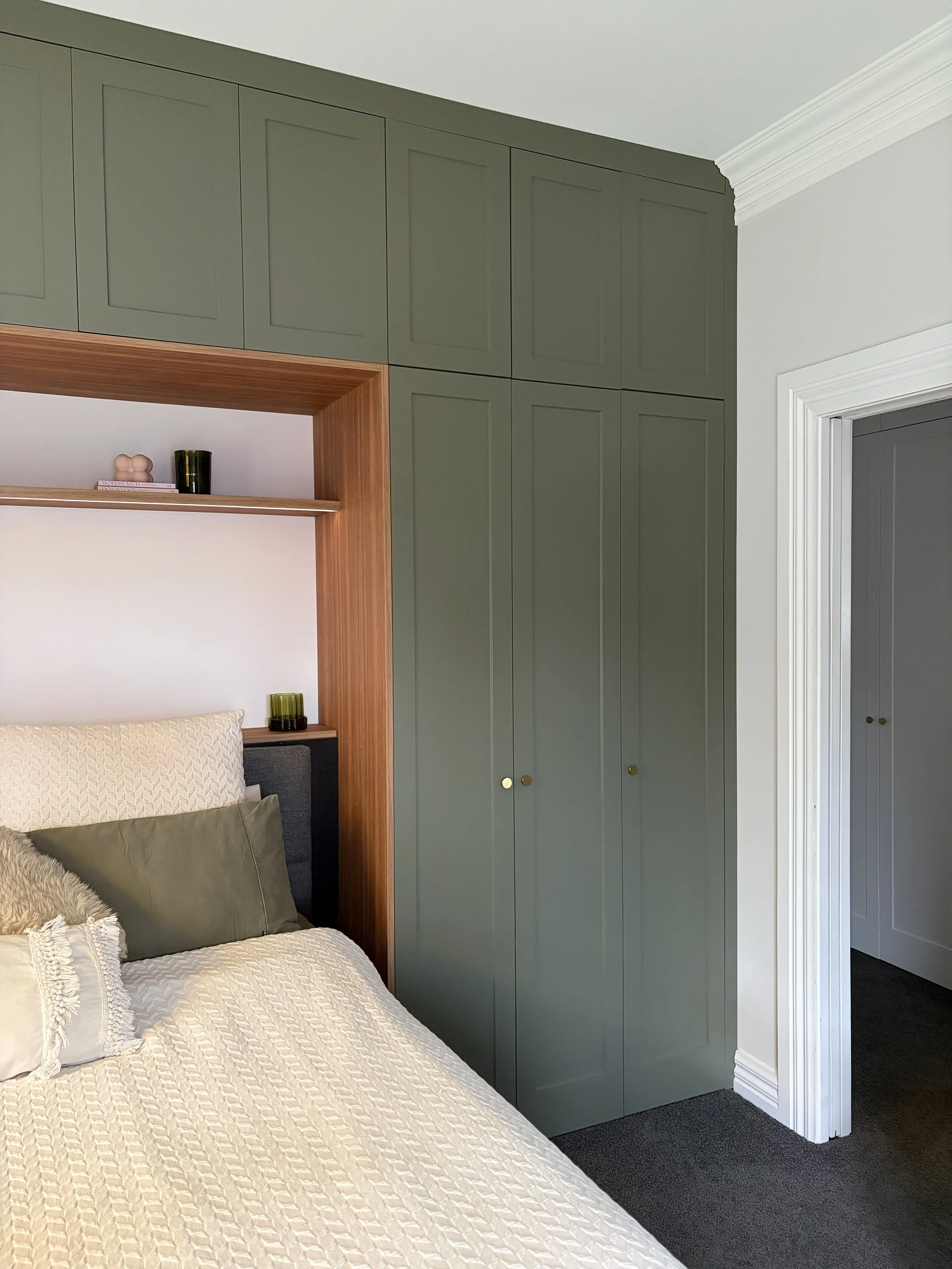 Close-up of a bedroom corner showing a bed with pillows and a built-in wardrobe in muted green with small round gold handles.