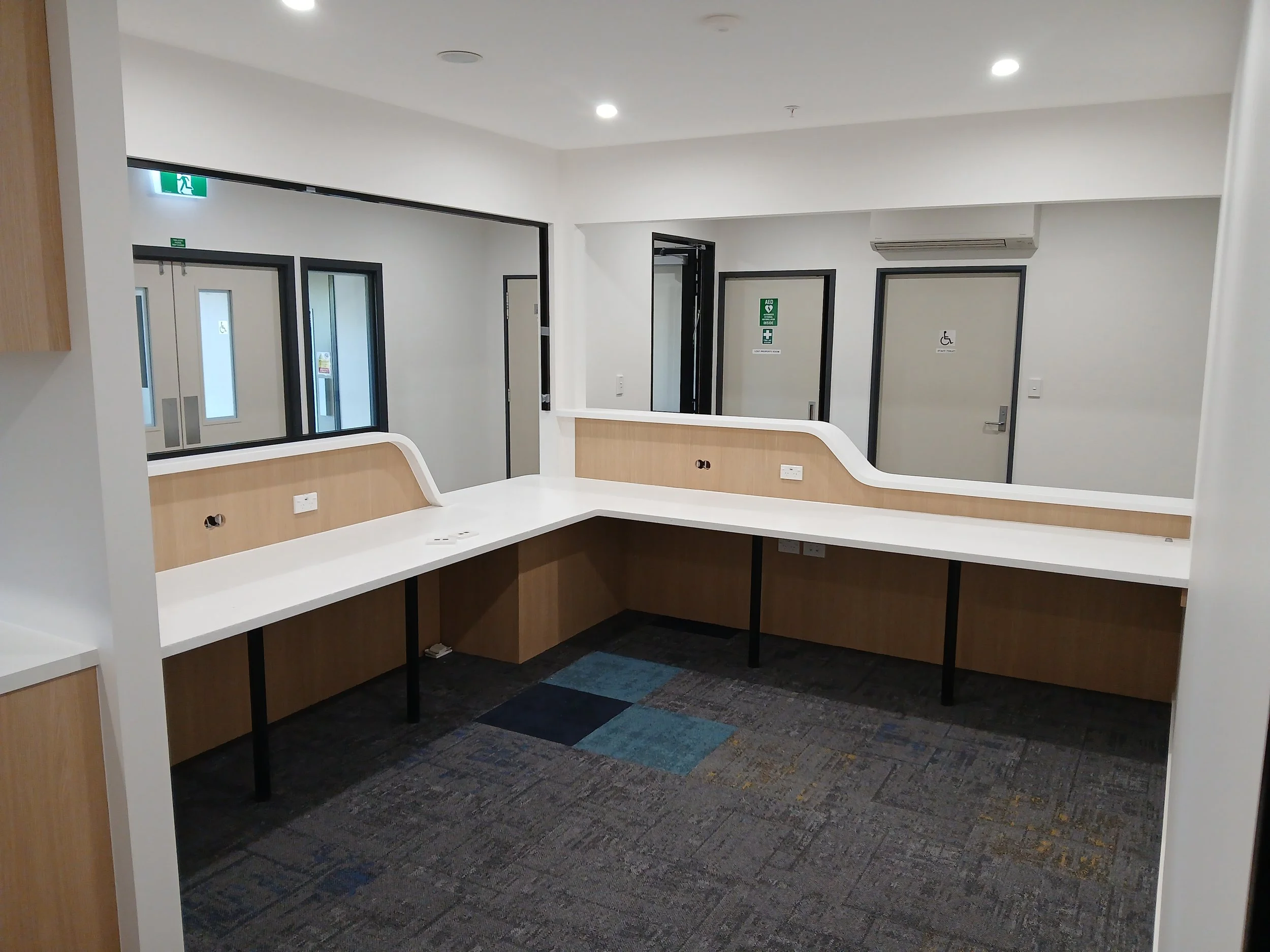 Empty office or reception area with white countertops, light wood paneling, black supports, and three doors with disability and emergency exit signs, carpeted floor, and an air conditioning unit on the wall.