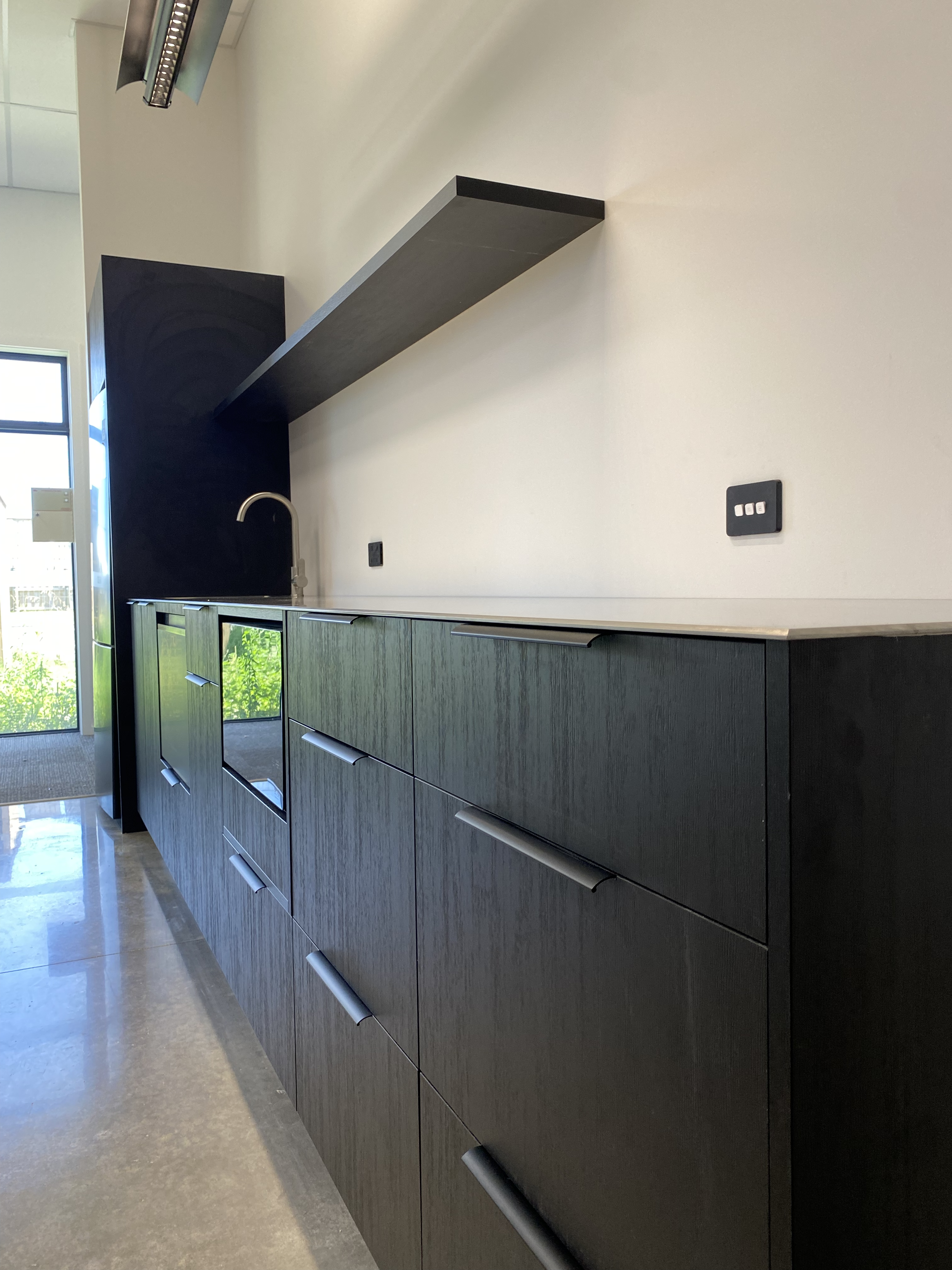 Modern black kitchen cabinet with silver handles, a white countertop, a black backsplash, a chrome faucet, a floating black shelf, and a large window showing greenery outside. Modern staff kitchen in the break room, easy access and functional flow.