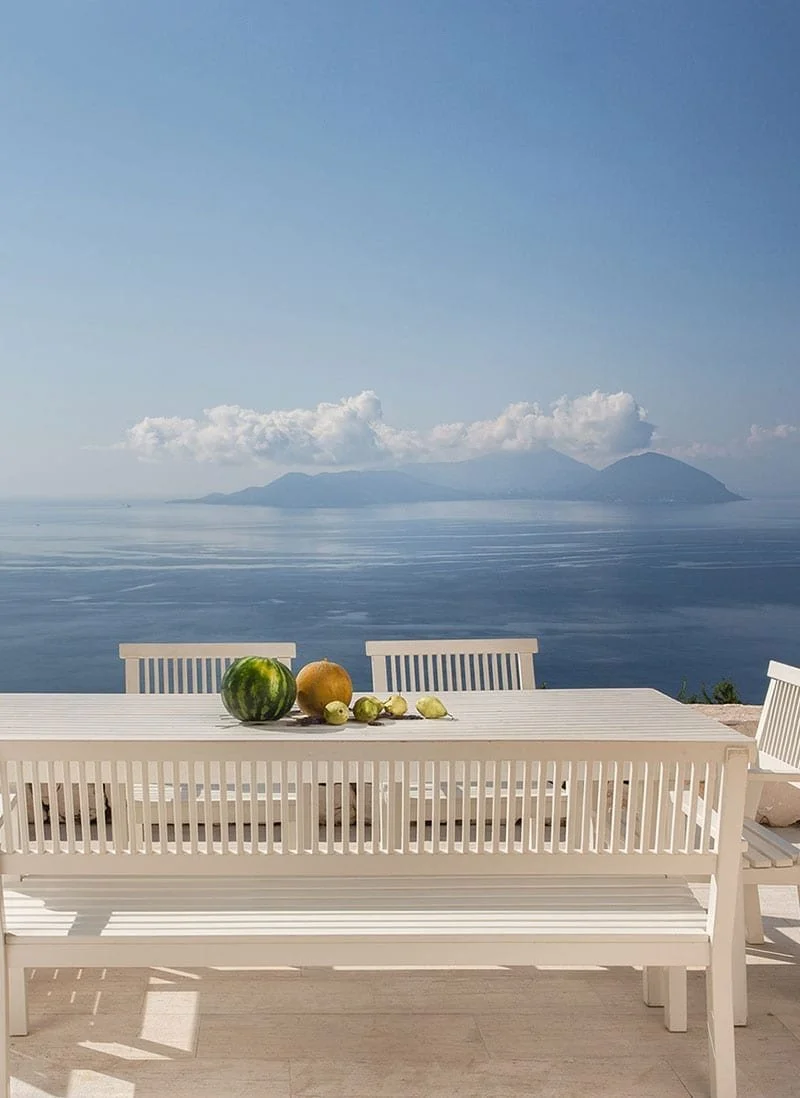 A white outdoor dining table with chairs overlooking a body of water and distant mountains, with a watermelon, melon, and grapes on the table.