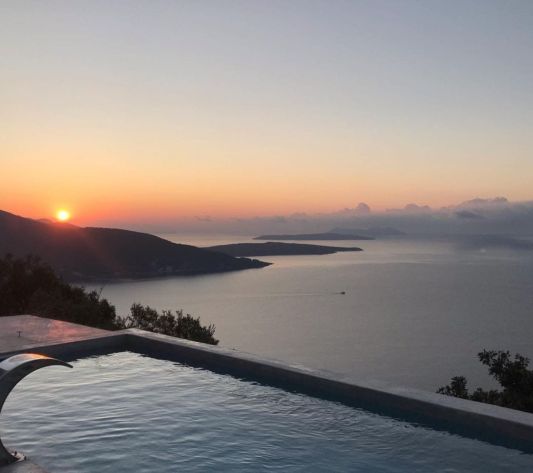 Sunset over a body of water viewed from a deck with an infinity pool, with hills and distant islands in the background.