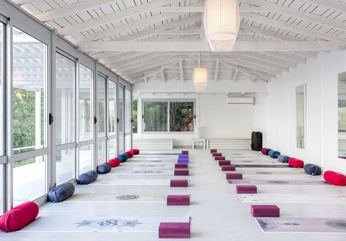 A bright yoga studio with wooden floors and white walls, featuring large glass windows on one side, yoga mats with blocks and bolsters, and pendant lights hanging from the vaulted ceiling.