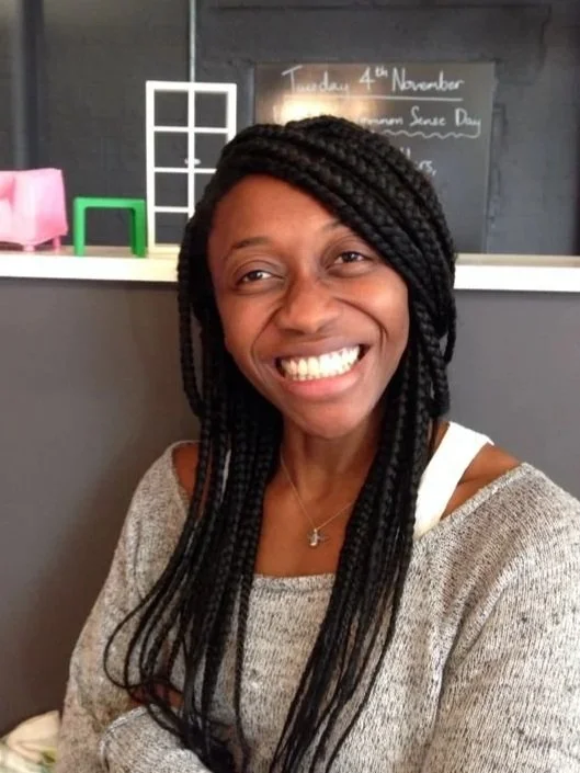 Helen Bazuaye, a professional coach, smiling with long braided hair wearing a grey sweater in a cafe.