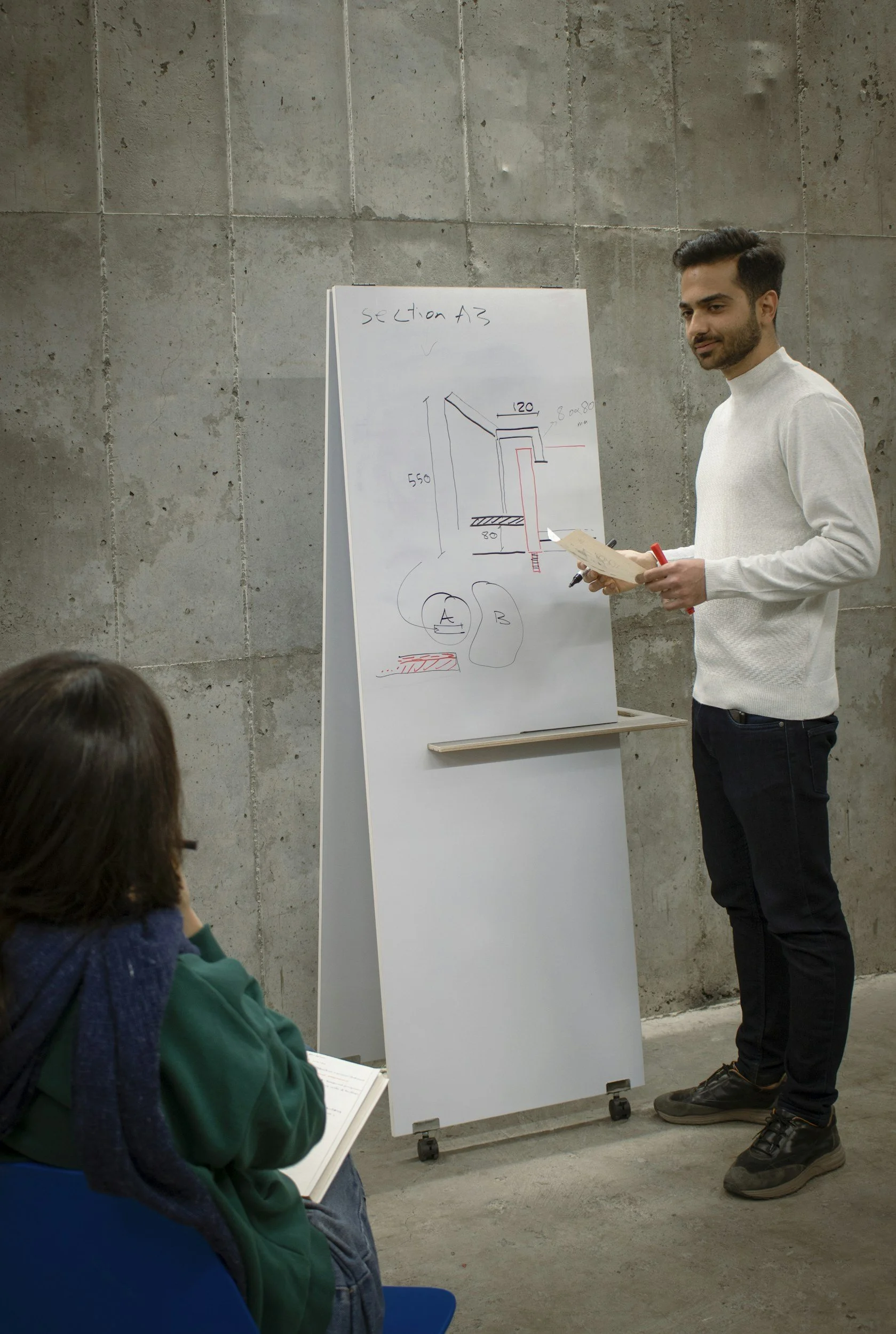 A man standing next to a whiteboard with technical drawings, addressing a seated person taking notes in a conference or classroom setting.