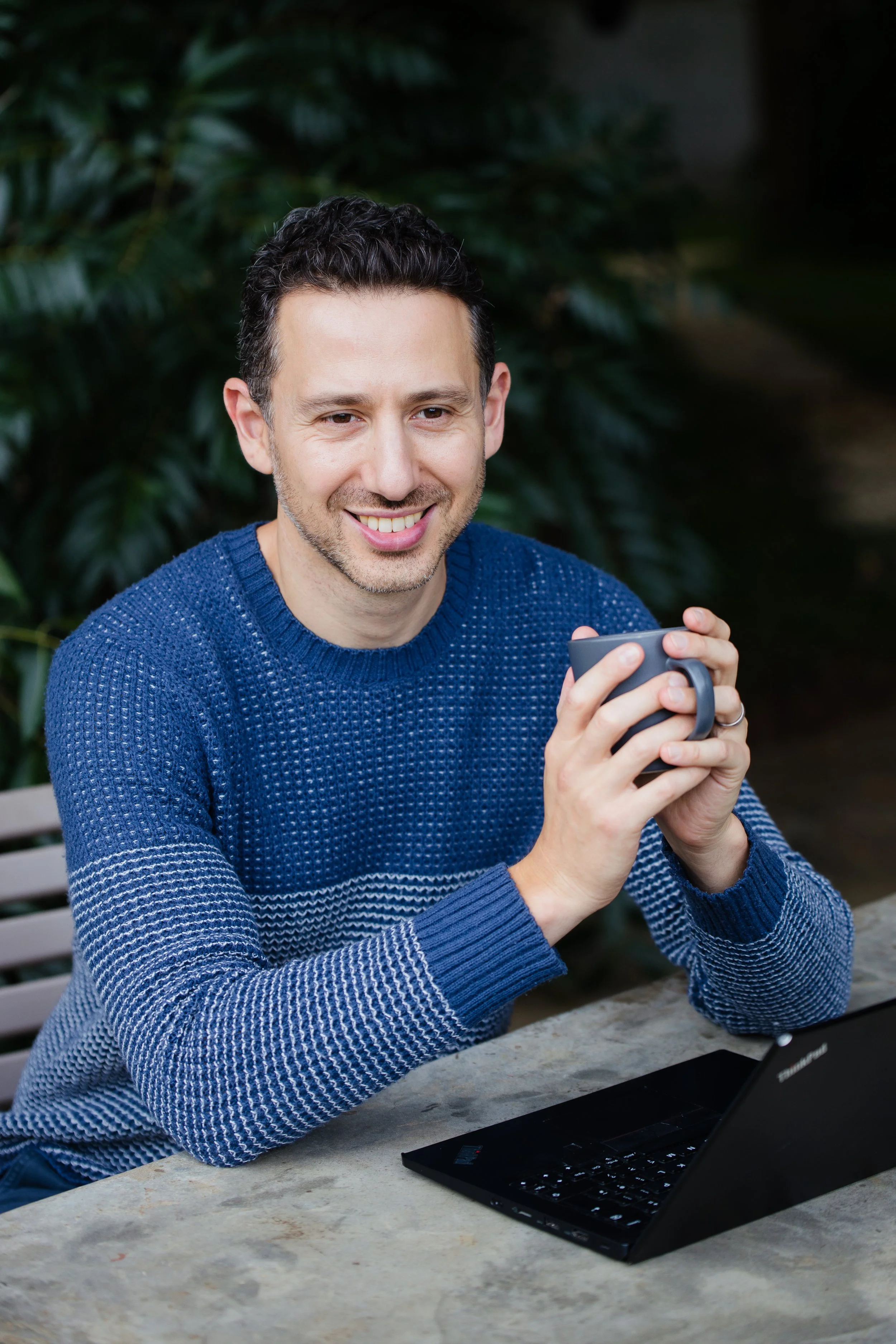 A man with dark, curly hair and a beard, wearing a blue sweater, smiling and holding a smartphone while sitting at a table with a laptop and a tablet in front of him. There are green plants in the background.