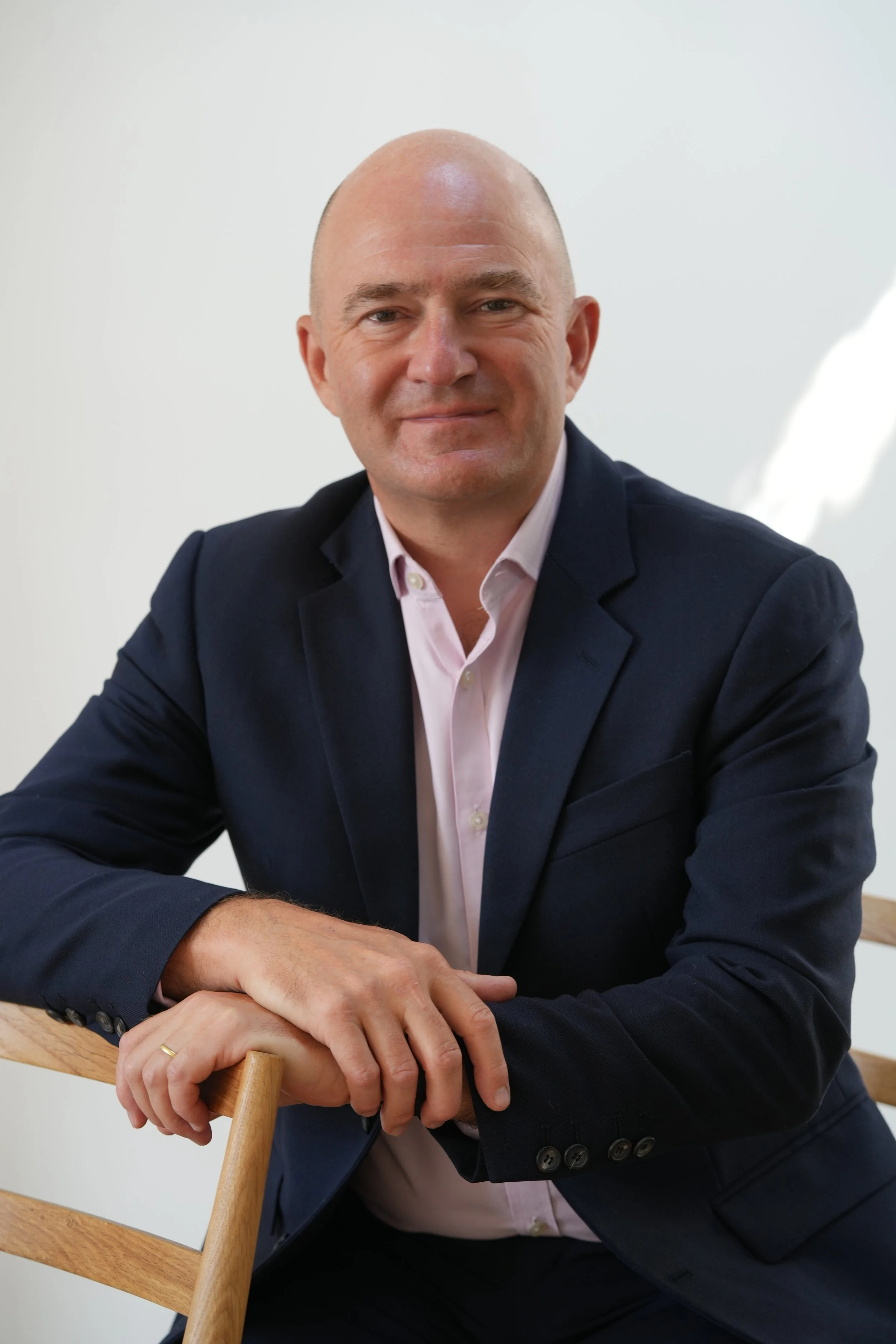 A smiling middle-aged man with a bald head, dressed in a dark suit, white shirt, and pink tie, against a plain white background.