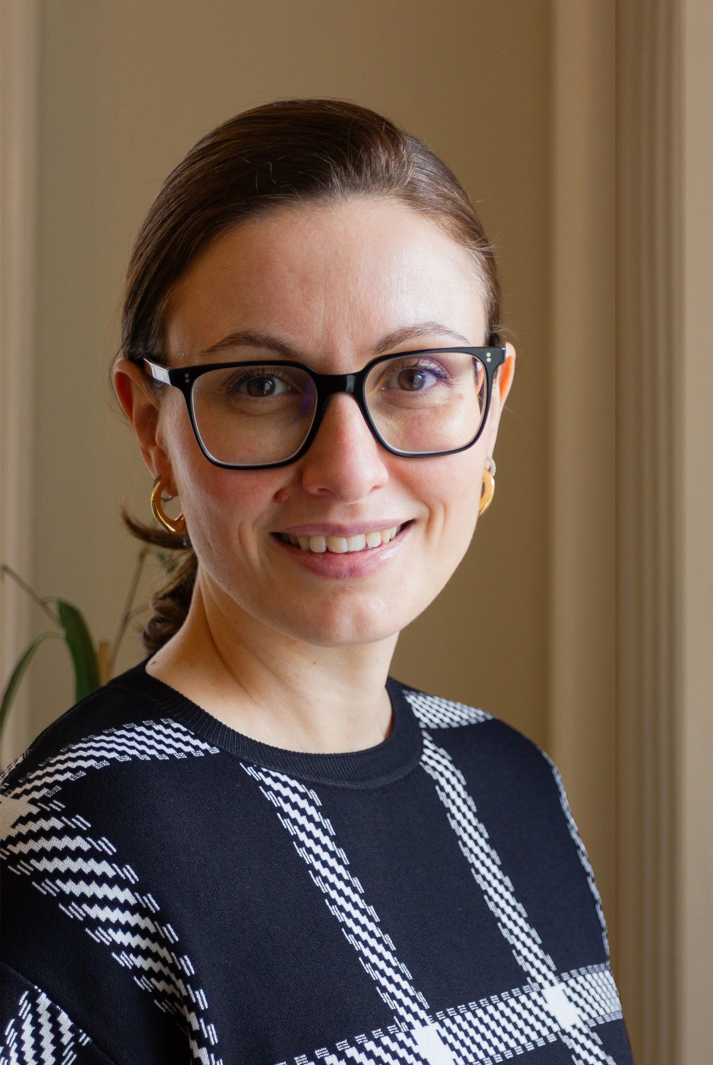 A smiling woman with glasses, wearing a black and white patterned sweater, with earrings, in an indoor setting.