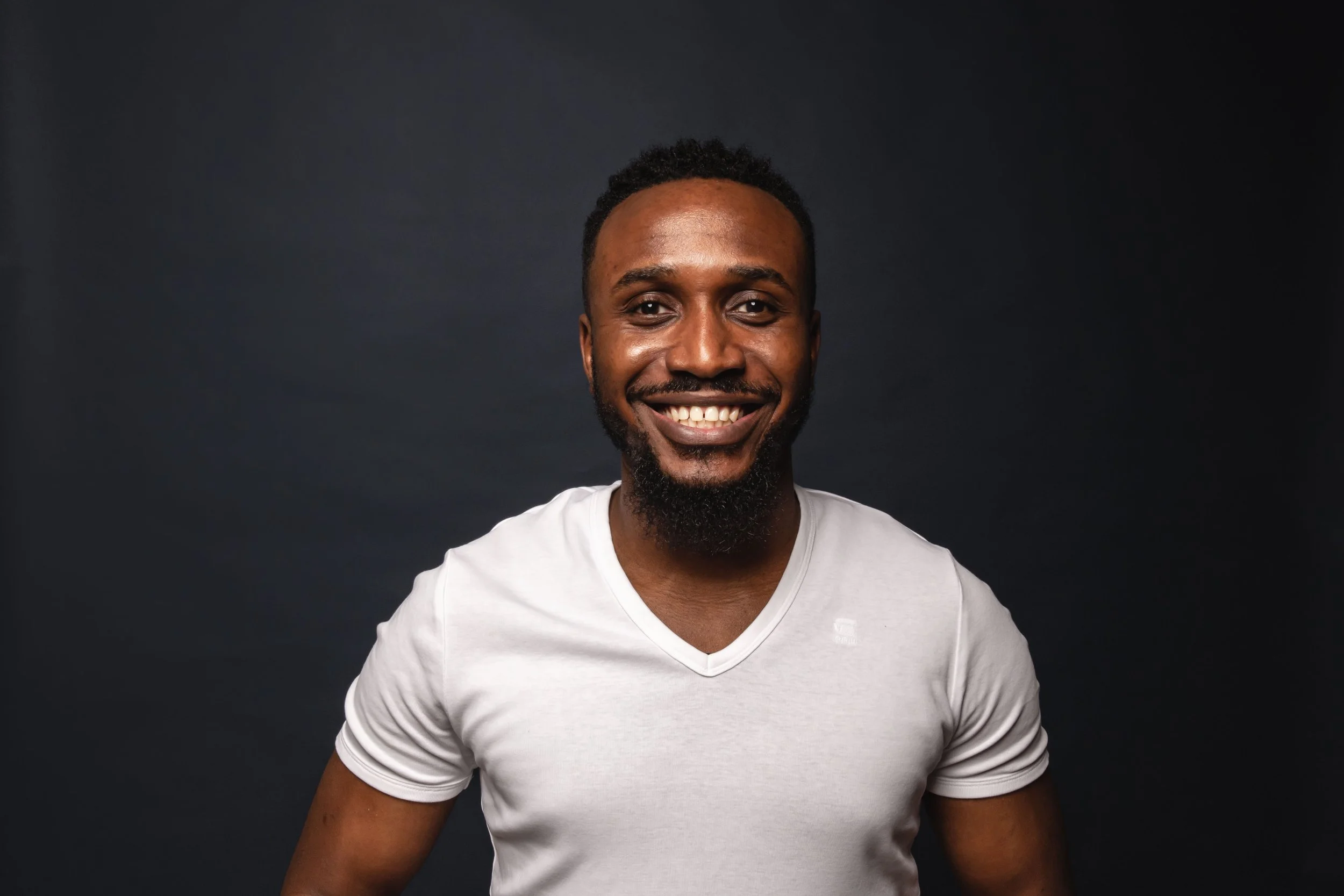 A smiling African American man with a beard and curly hair, wearing a white T-shirt, standing against a dark background.