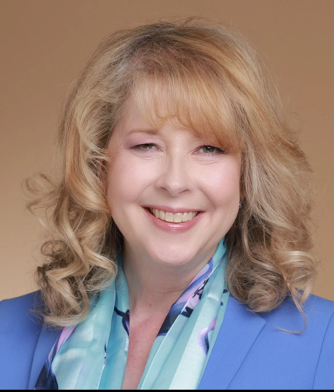 Portrait of a middle-aged woman with blonde curly hair smiling with closed lips, wearing a blue blazer and a colorful scarf, against a plain background.