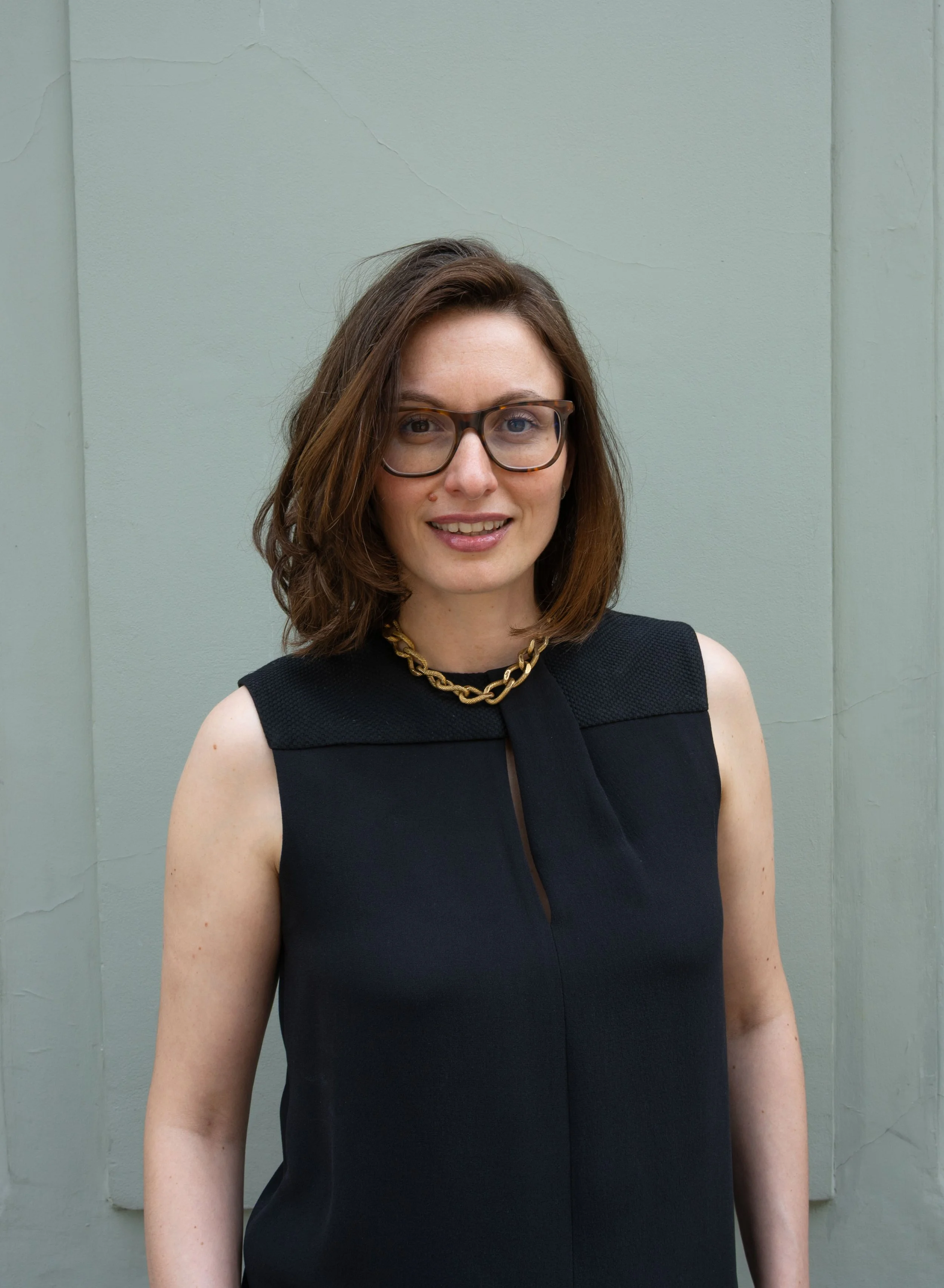 Portrait of a woman with shoulder-length brown hair, wearing glasses, a black sleeveless top, a gold chain necklace, and standing in front of a light green wall with visible cracks.