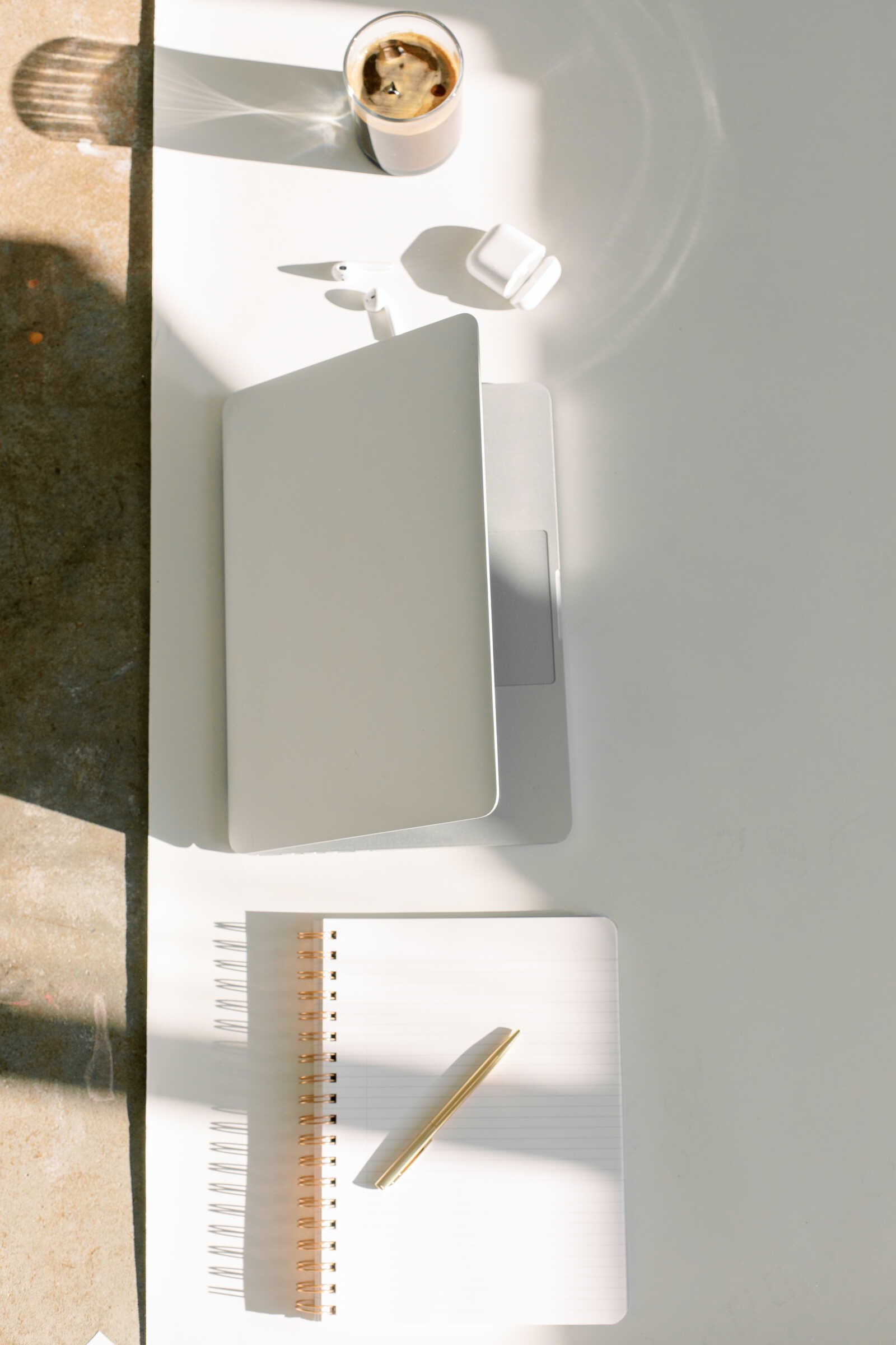 Kuvassa on auki oleva kannettava tietokone, parit langattomat kuulokkeet, älypuhelin, muistikirja ja kyna sekä lasi kahvia, jotka on asetettu valkoiselle pöydälle auringonvalossa.