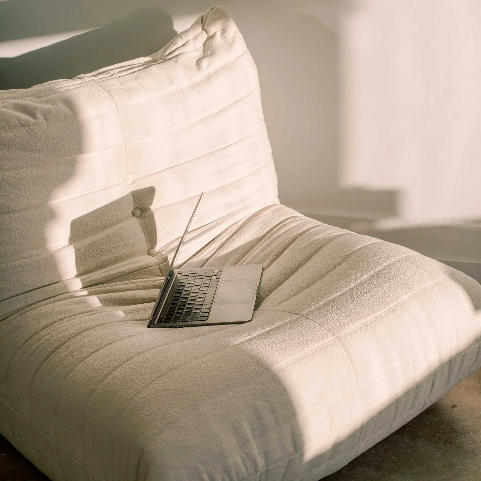 Vaalea togo-nojatuoli, jossa on avattu pieni kannettava tietokone, ja huoneeseen laskeva auringonvalo luo varjon.