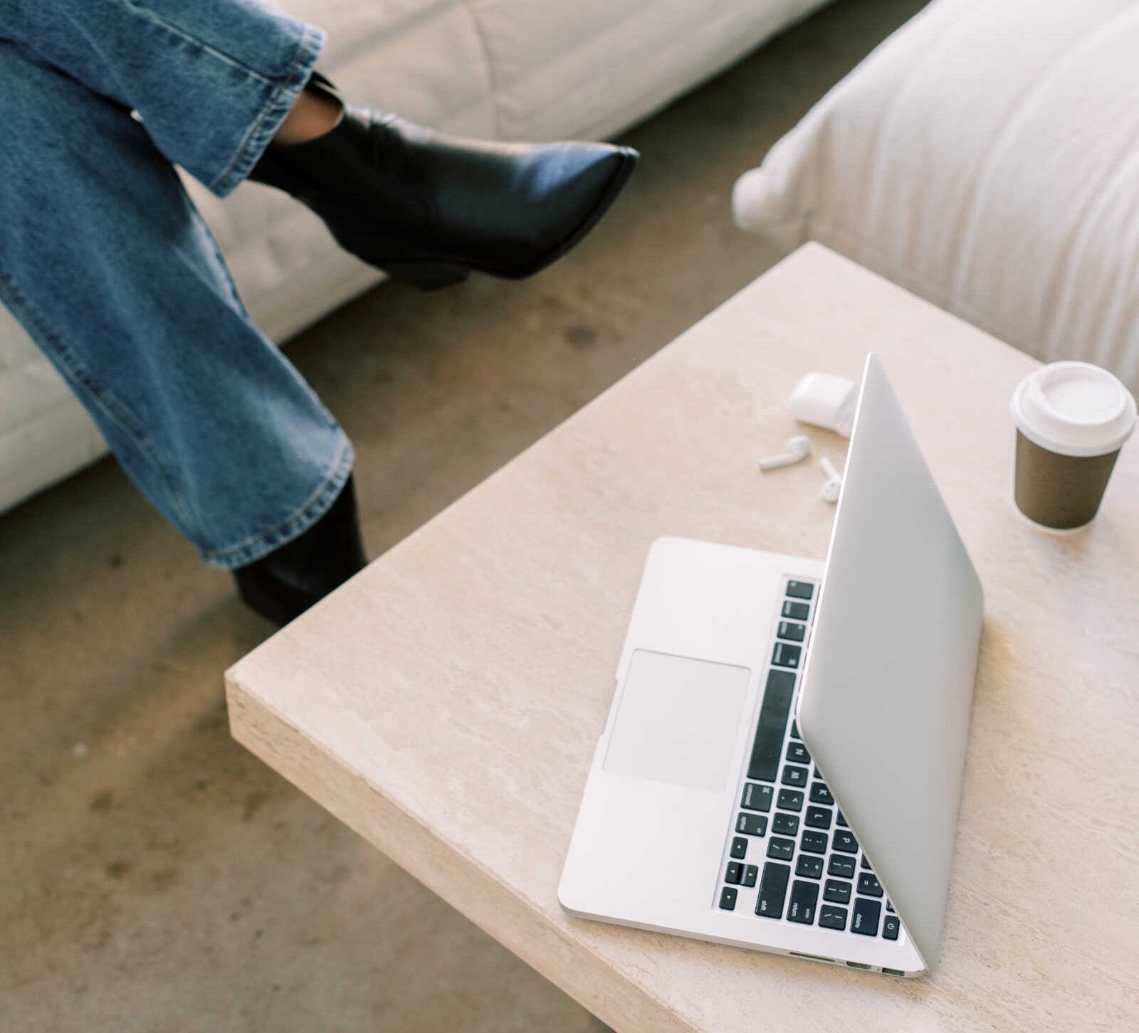 Nainen istuu sohvalla jalat ristissä, päällä farkut ja nilkkakorkokengät, työpöydällä on avattu tietokone, kahvikuppi ja lääkkeitä.