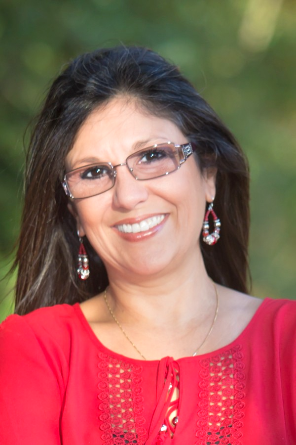 A smiling woman with glasses and earrings, wearing a red top, outdoors with a blurred green background. Sandy McDonald, CEO Digitally Driven Solutions, and Things to do at cedar creek lake.
