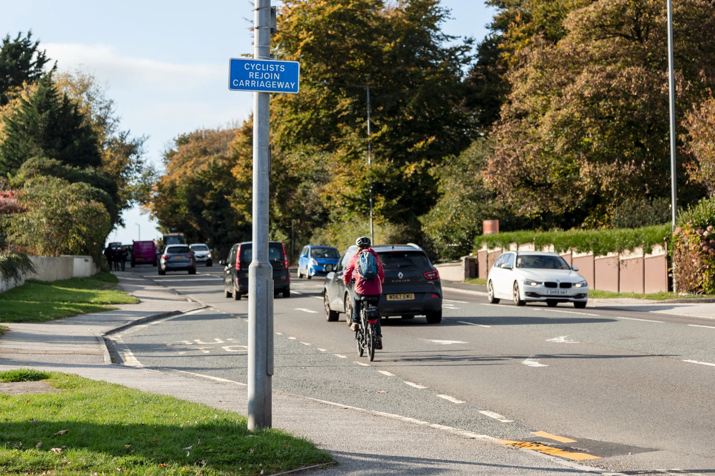 A cyclist riding on a bike lane on a city street with cars, trees with fall foliage, and a blue road sign indicating a shared space for cyclists and vehicle rejoin at a carriageway.