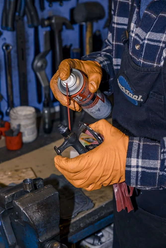 A person wearing orange gloves and a plaid shirt applies lubricant to a bicycle component, possibly a shifter or derailleur, in a workshop with tools hanging in the background.