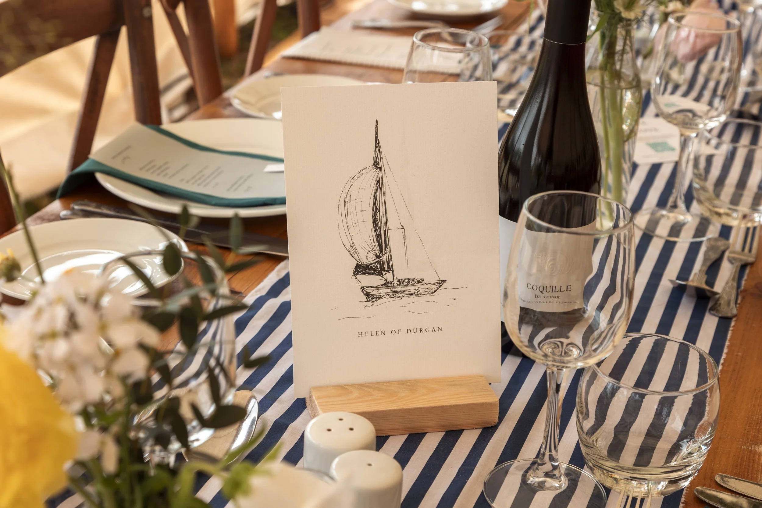 A table setting with a boat-themed place card featuring a sketch of a sailboat and the name Helen of Durgan, surrounded by wine glasses, plates, a black bottle, and a flower arrangement, with a blue and white striped table runner.