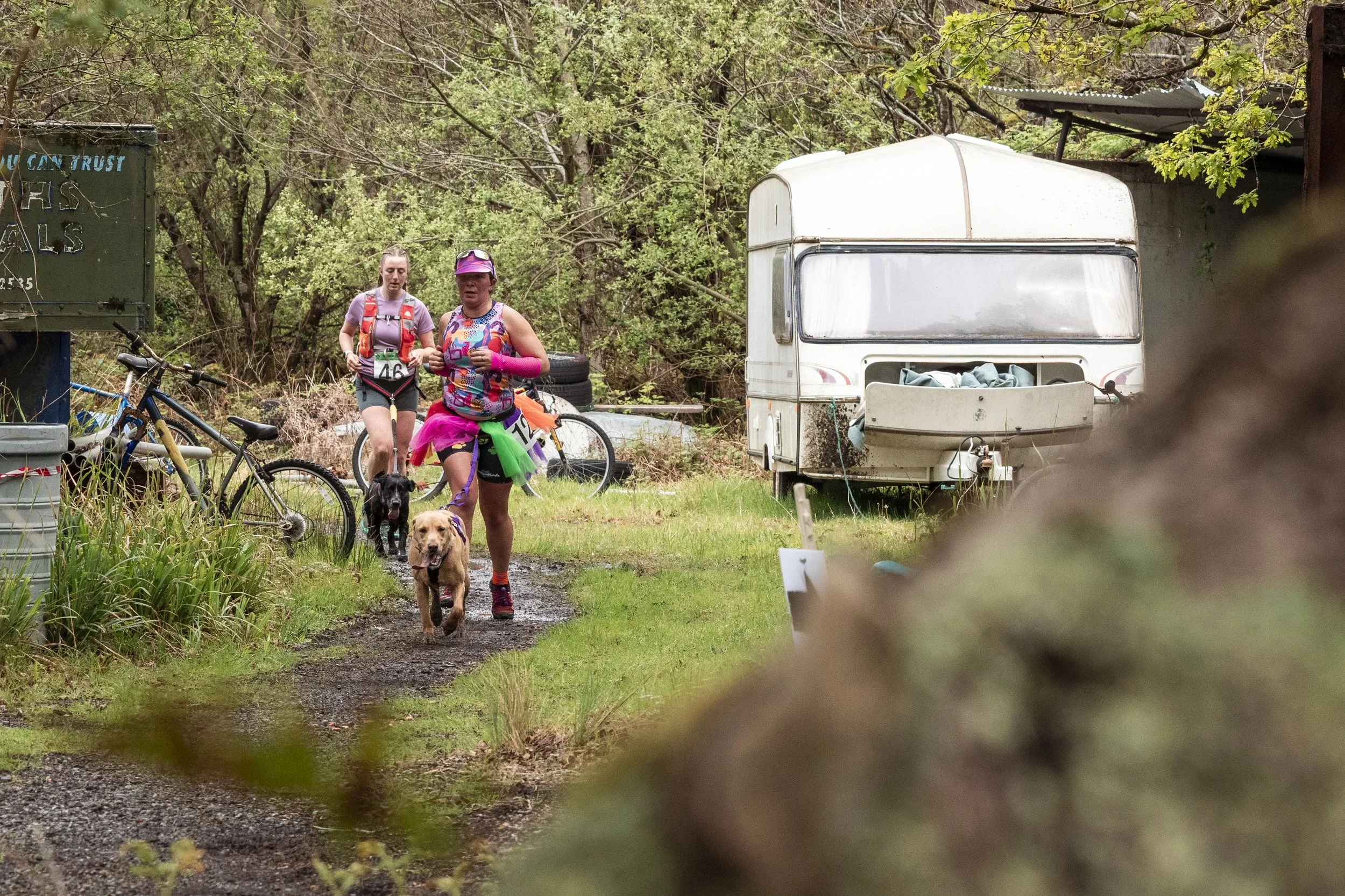Two women walking dogs with bicycles on a muddy trail near a camper trailer, surrounded by trees and greenery.