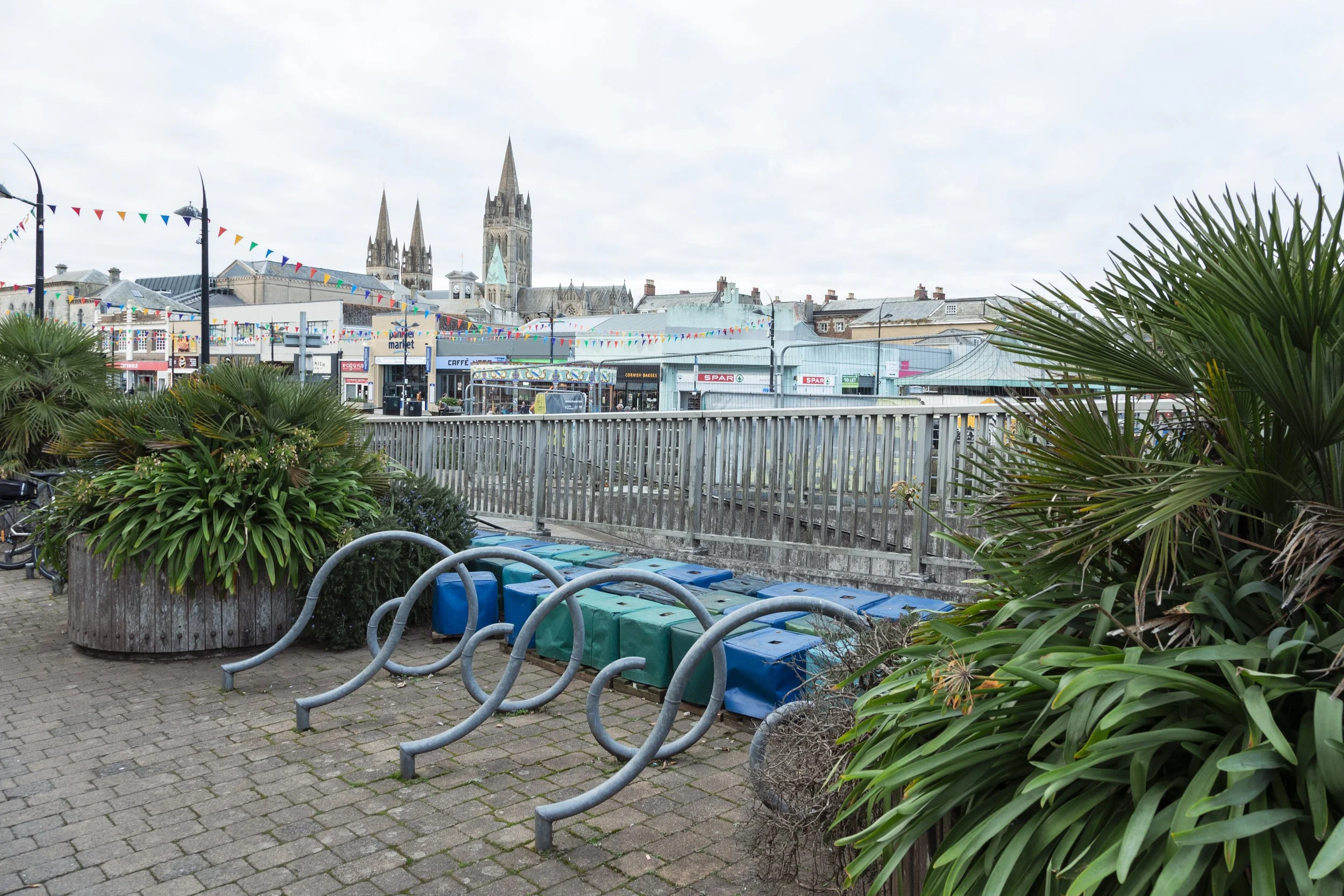 Empty bike racks and large potted plants on a paved area, with a fence and a street scene with shops, bunting decorations, and a church with spires in the background under an overcast sky.