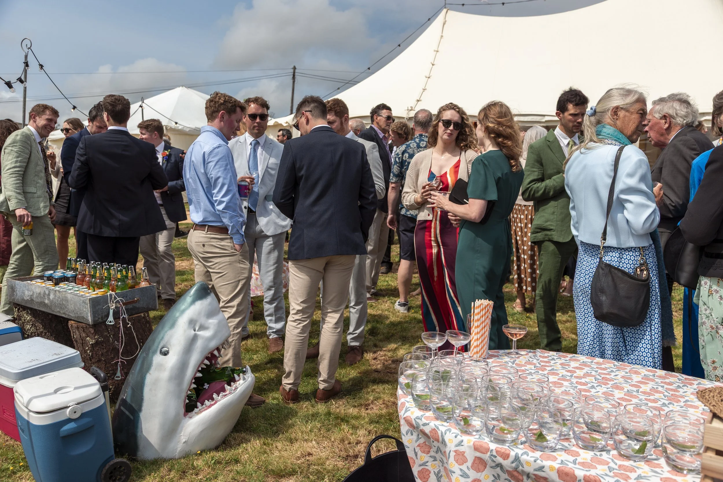 People socializing at an outdoor party under a tent, with drinks and a table of glasses, and a shark head decoration on the ground.
