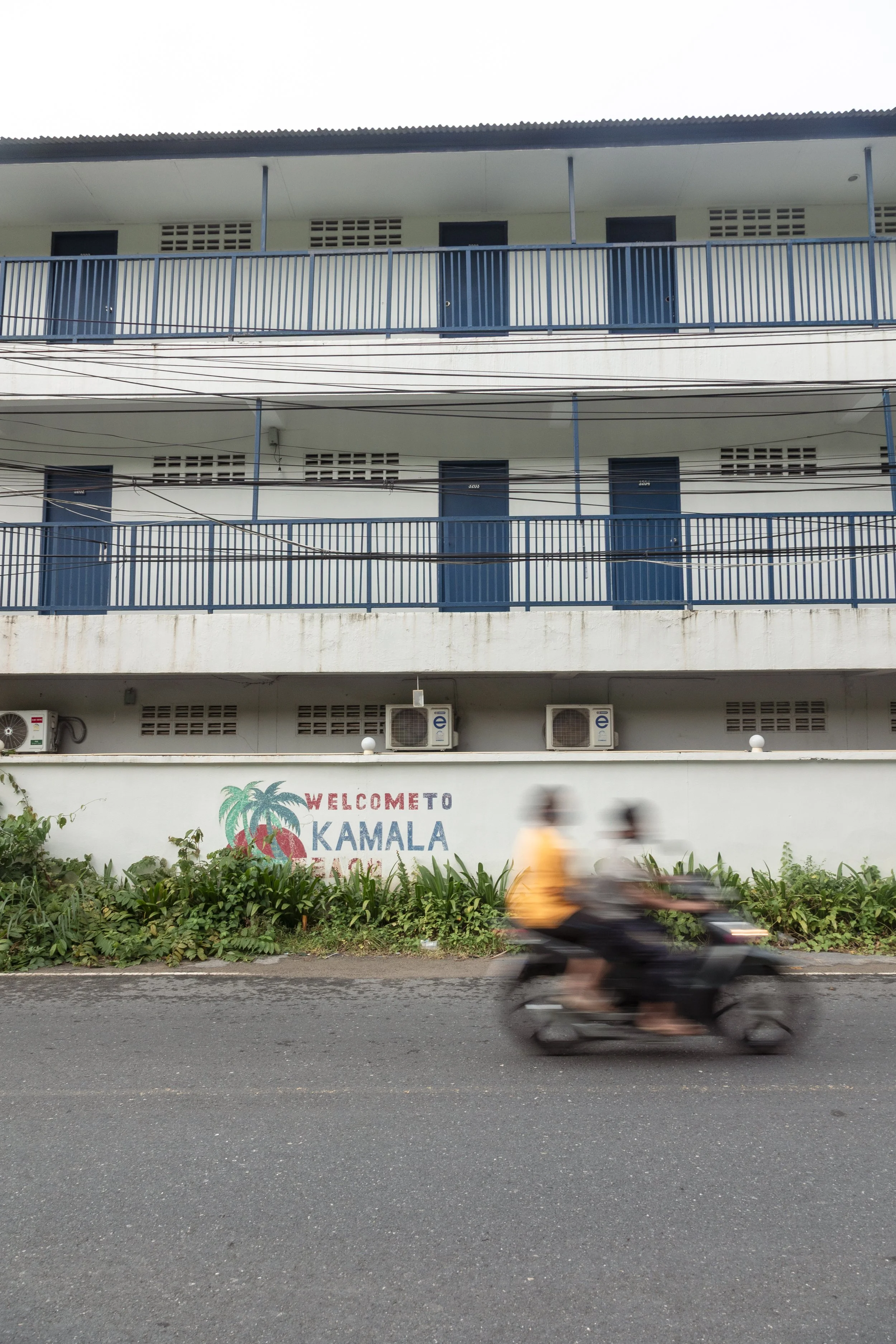 A building with blue balconies and air conditioning units, with a sign that says "Welcome to Kamala Beach" on the wall, and a motorcycle with two riders passing by in front of the building.