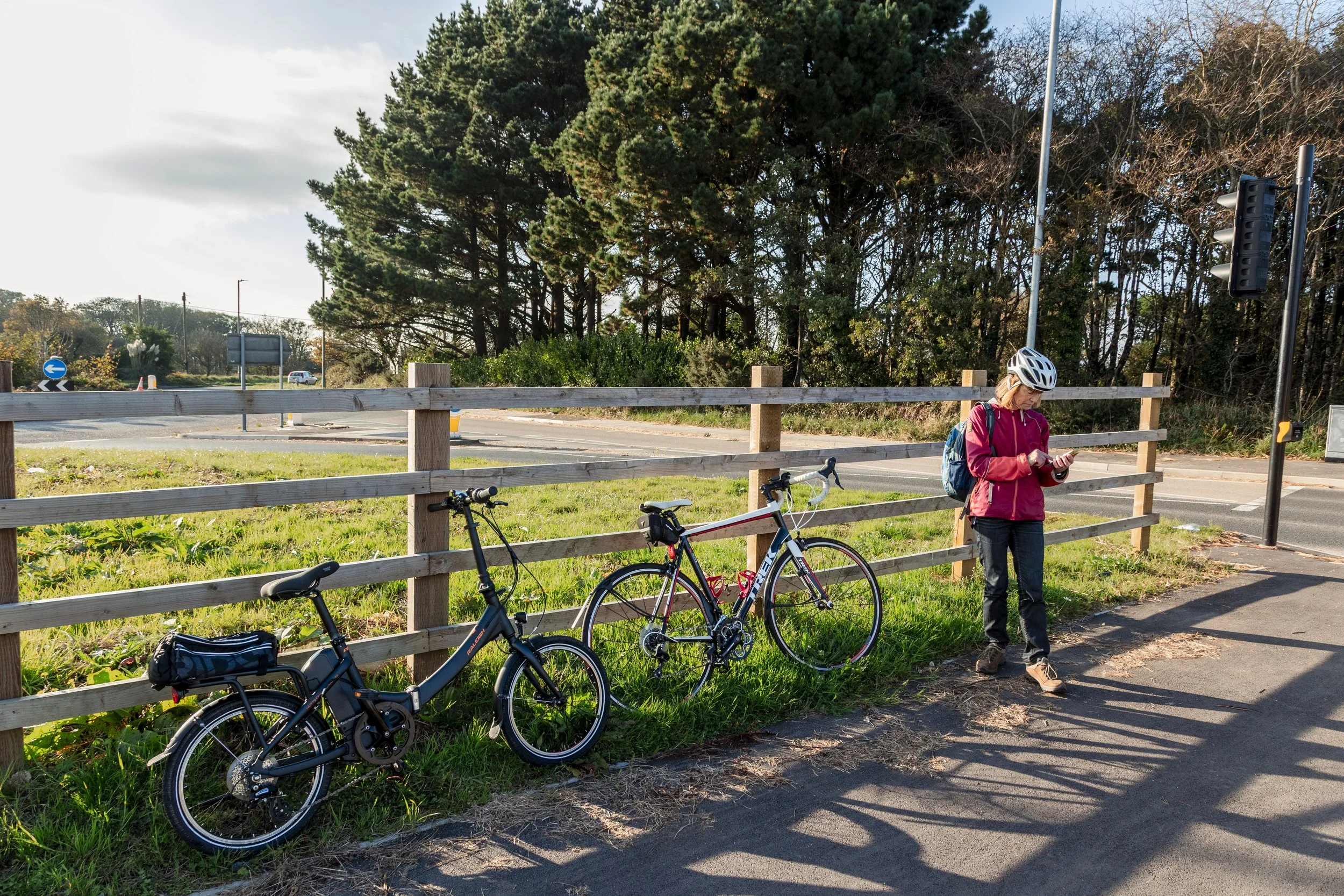 A woman in a red jacket, black pants, and a white helmet standing on a sidewalk, looking at her phone. Two bicycles, one black and one white with red accents, lean against a wooden fence behind her. There is green grass and trees in the background, a