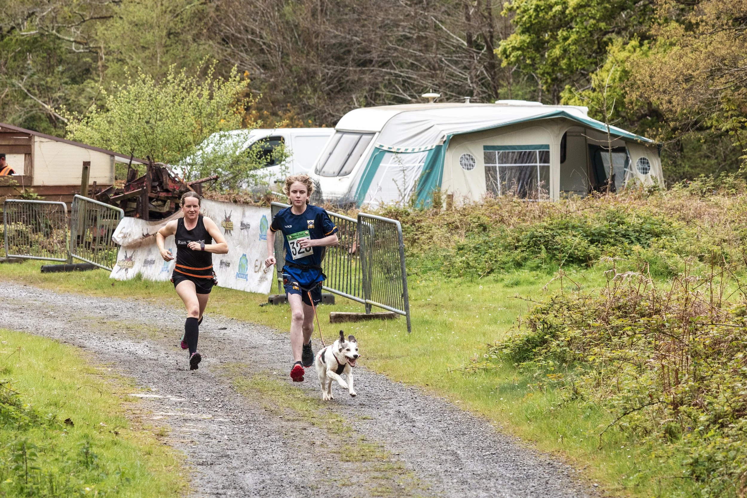 Two women running on a gravel trail with a dog in front of them. Behind them is a camping area with a camper, a white van, and a small wooden building, surrounded by trees and greenery.