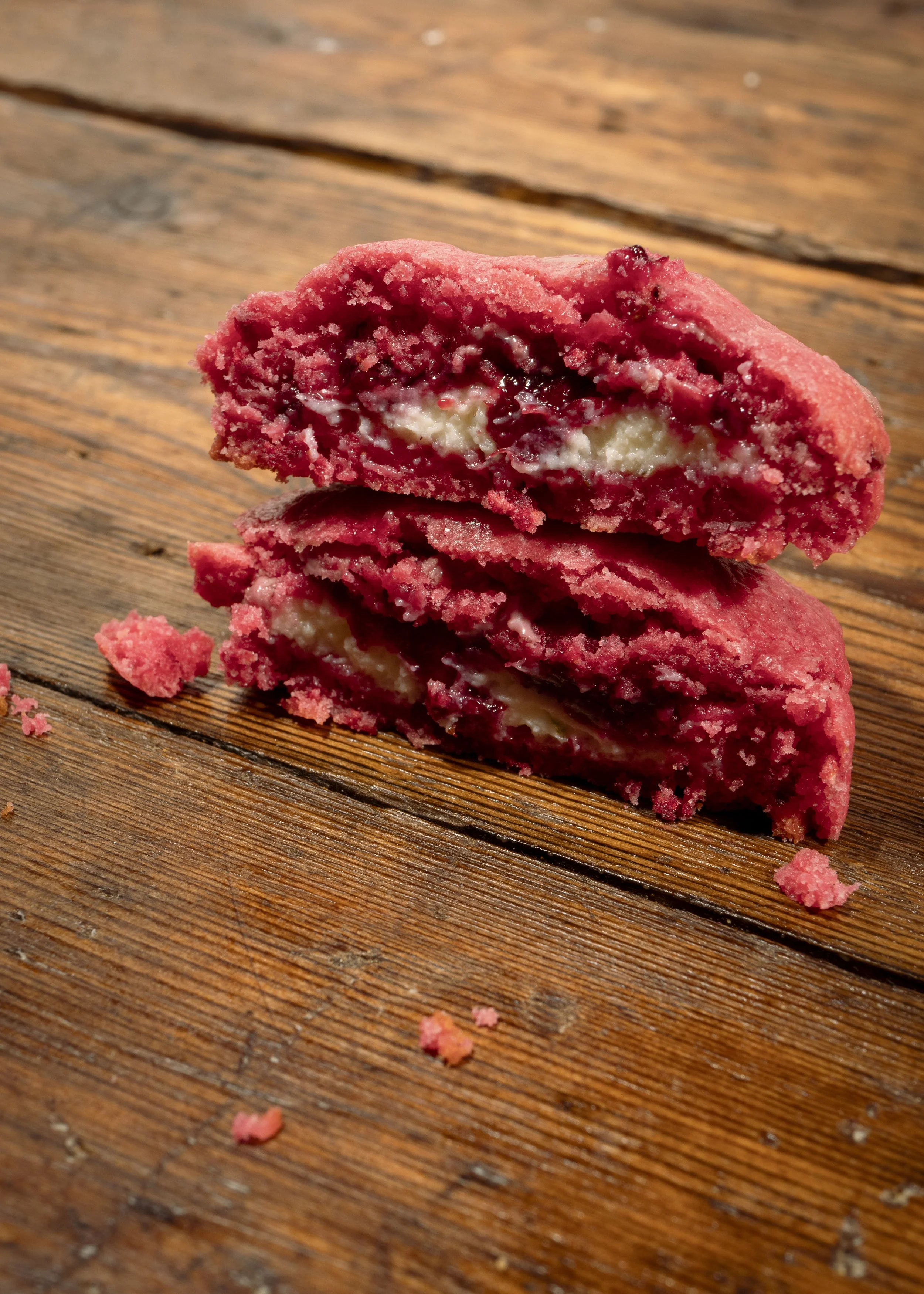A close-up of two stacked berry-flavored cookies with a creamy filling on a wooden surface, with crumbs scattered around.