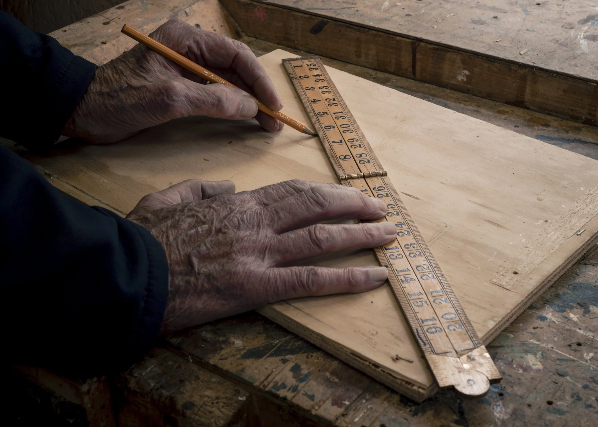 Older person marking a wooden piece with a pencil and measuring with a wooden ruler on a workbench.