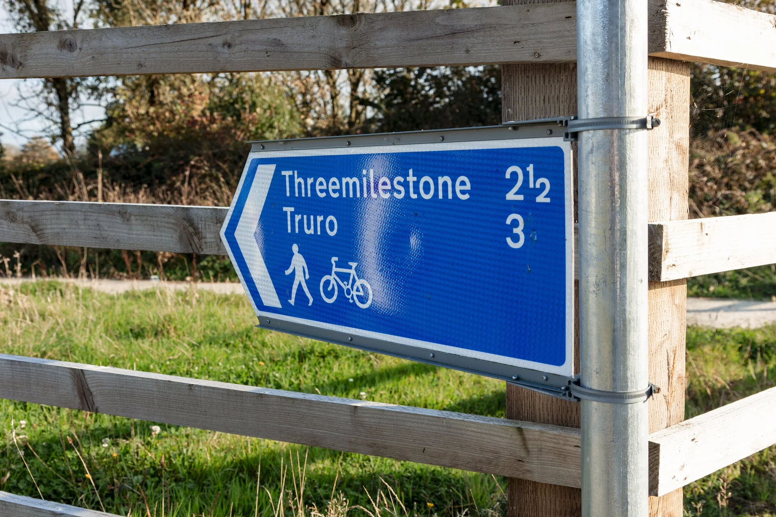 Blue directional sign with white text and symbols for walking and cycling, indicating distances to Threemilestone (2.5 miles) and Truro (3 miles), mounted on a wooden fence with greenery and trees in the background.