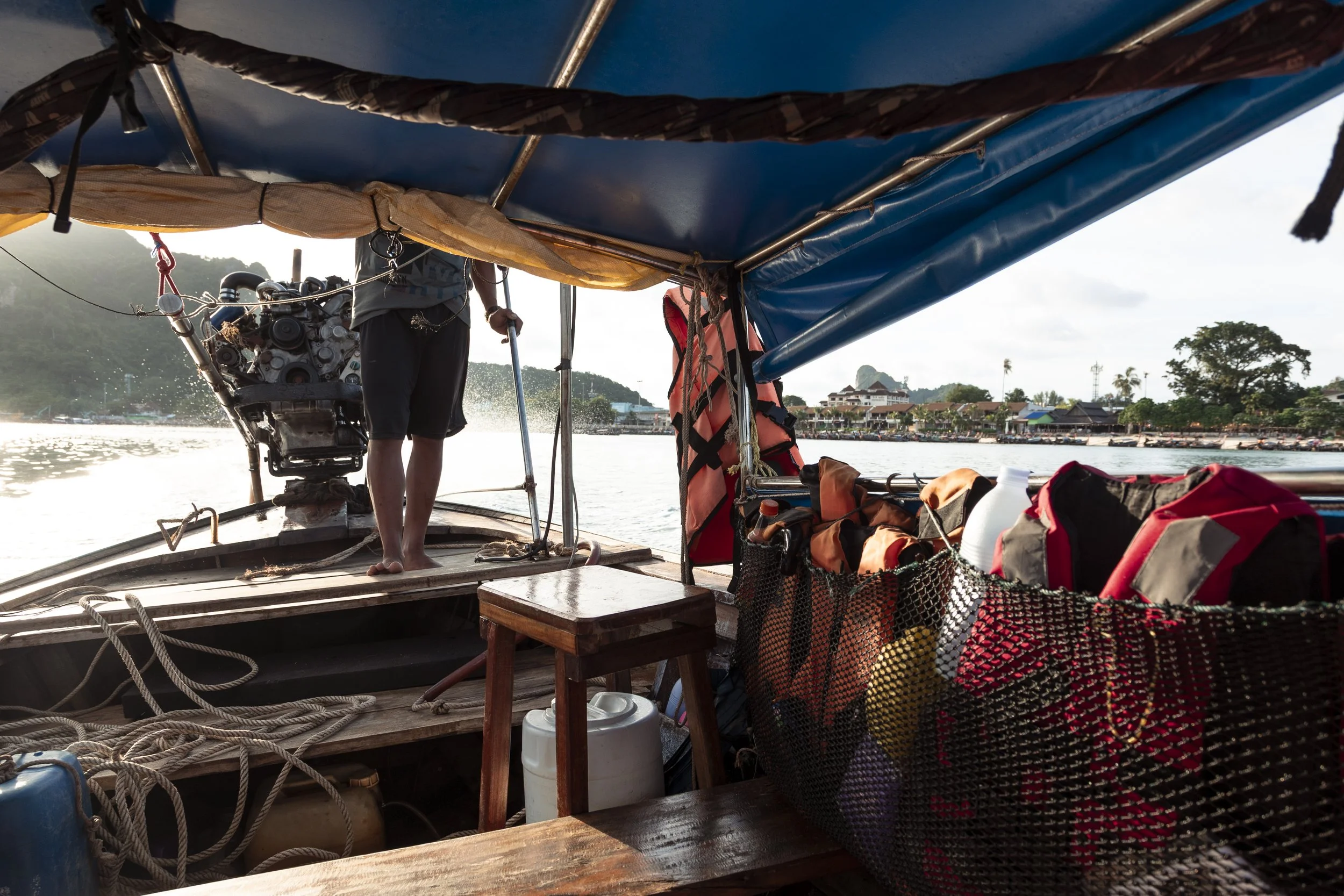 View from inside a boat looking toward the riverbank, showing a person working on an engine at the front of the boat, with luggage and life jackets stored inside.