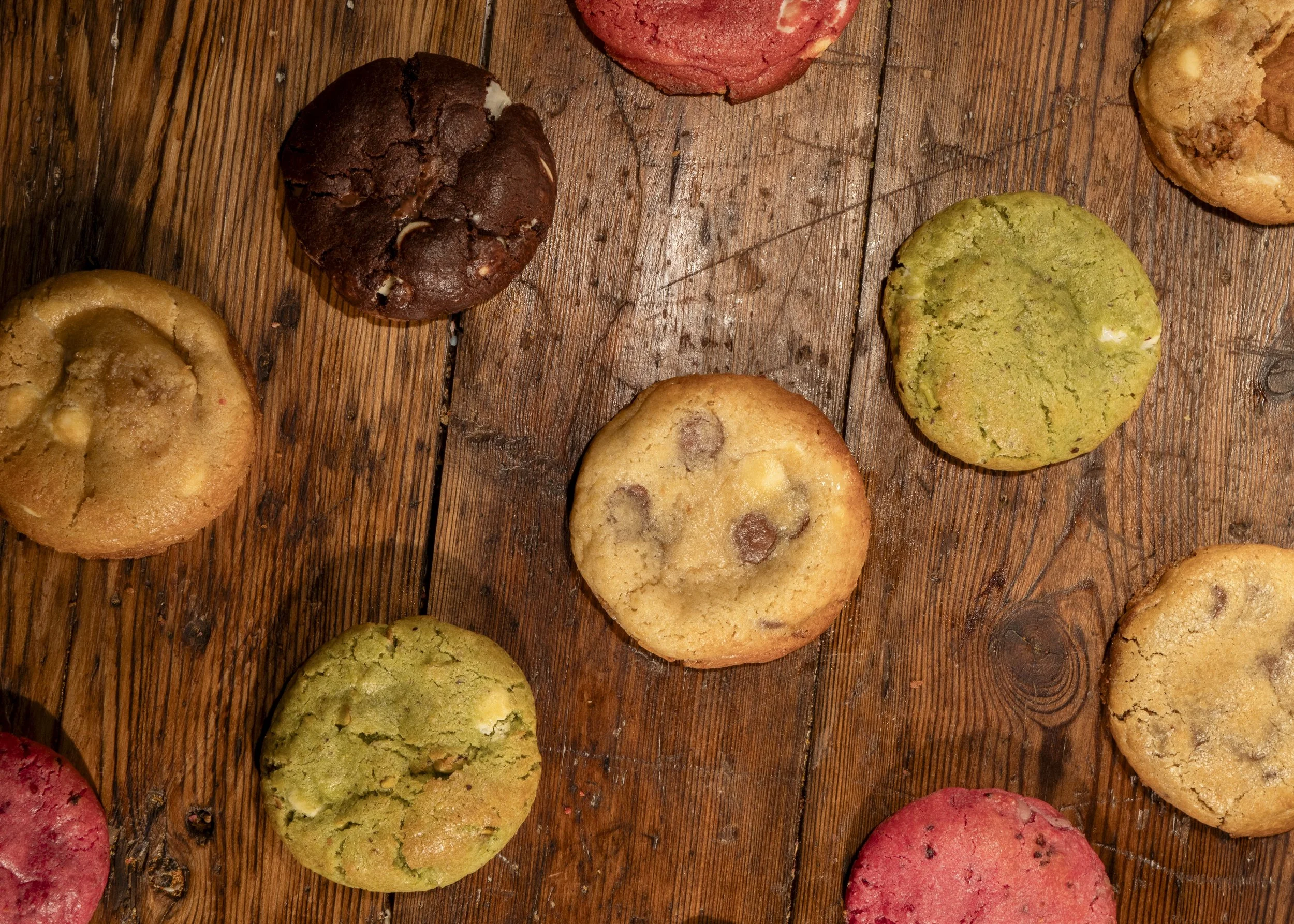 Assorted cookies on a wooden surface, including chocolate chip, green, red, and other flavored cookies.