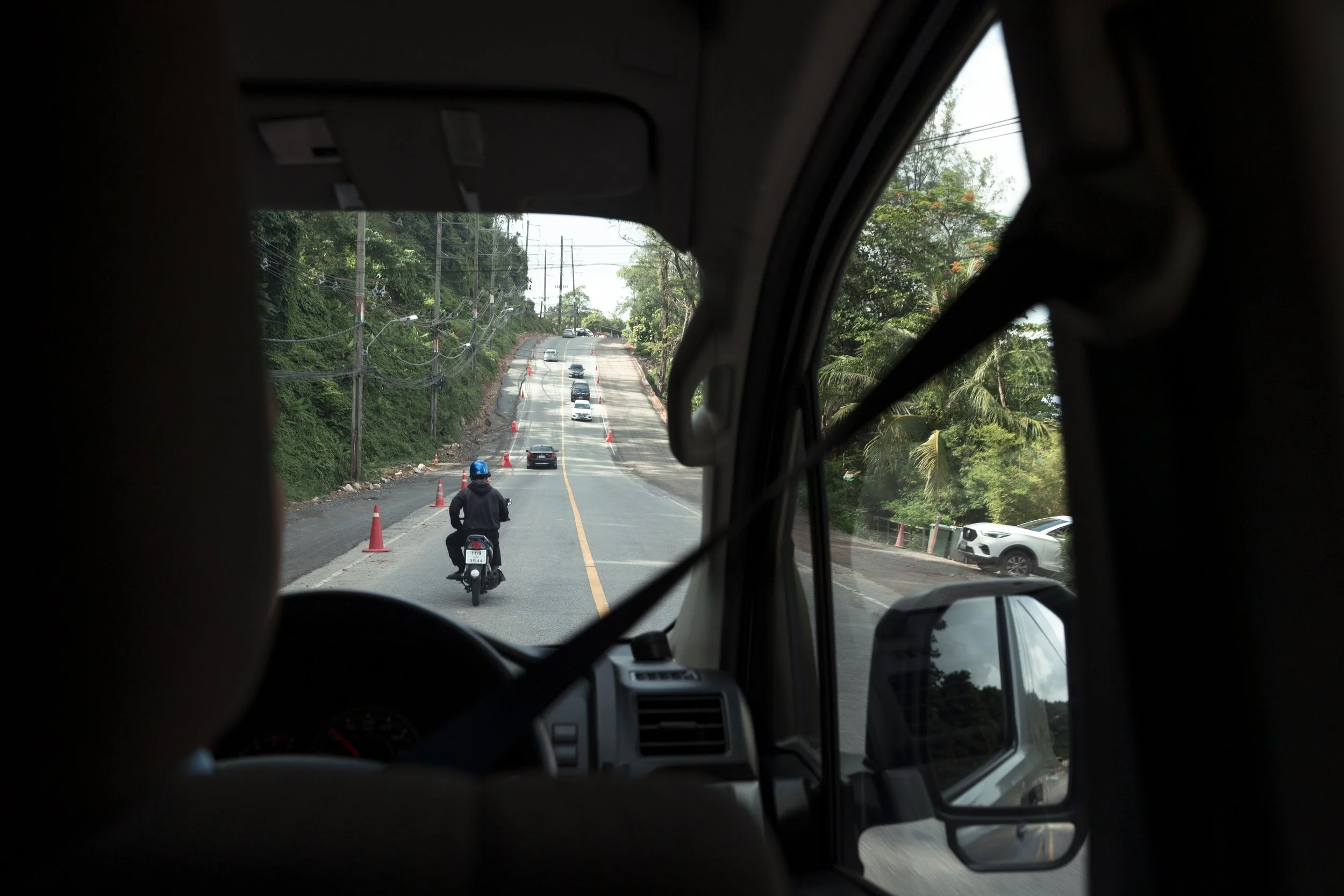 View from inside a vehicle showing a traffic checkpoint with cones and a motorcyclist passing through, on a road surrounded by greenery and trees.