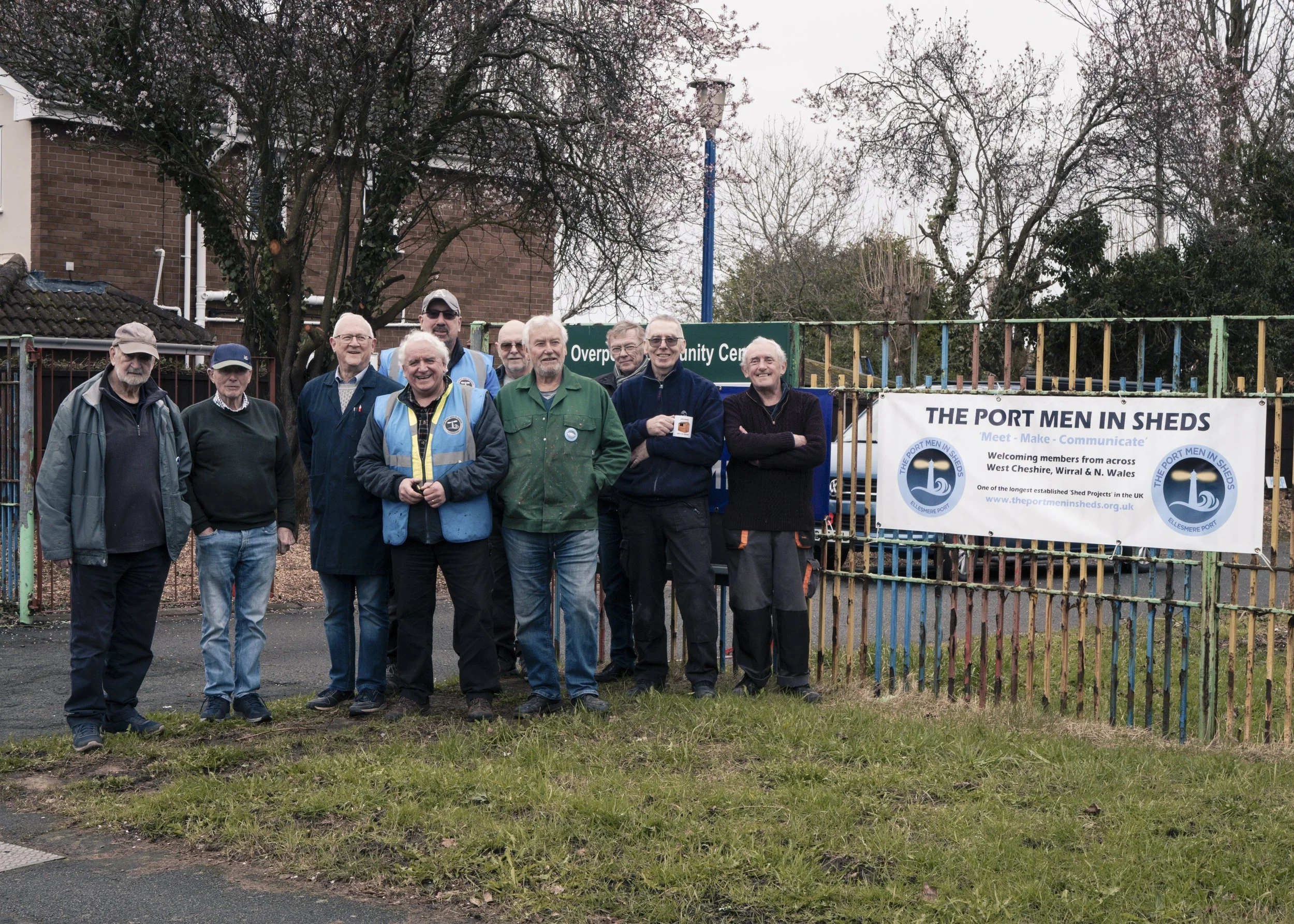 Group of men standing outdoors in front of a sign that reads 'The Port Men in Sheds' and 'Overpool Community Centre', with a colorful metal fence behind them, under leafless trees on a cloudy day.