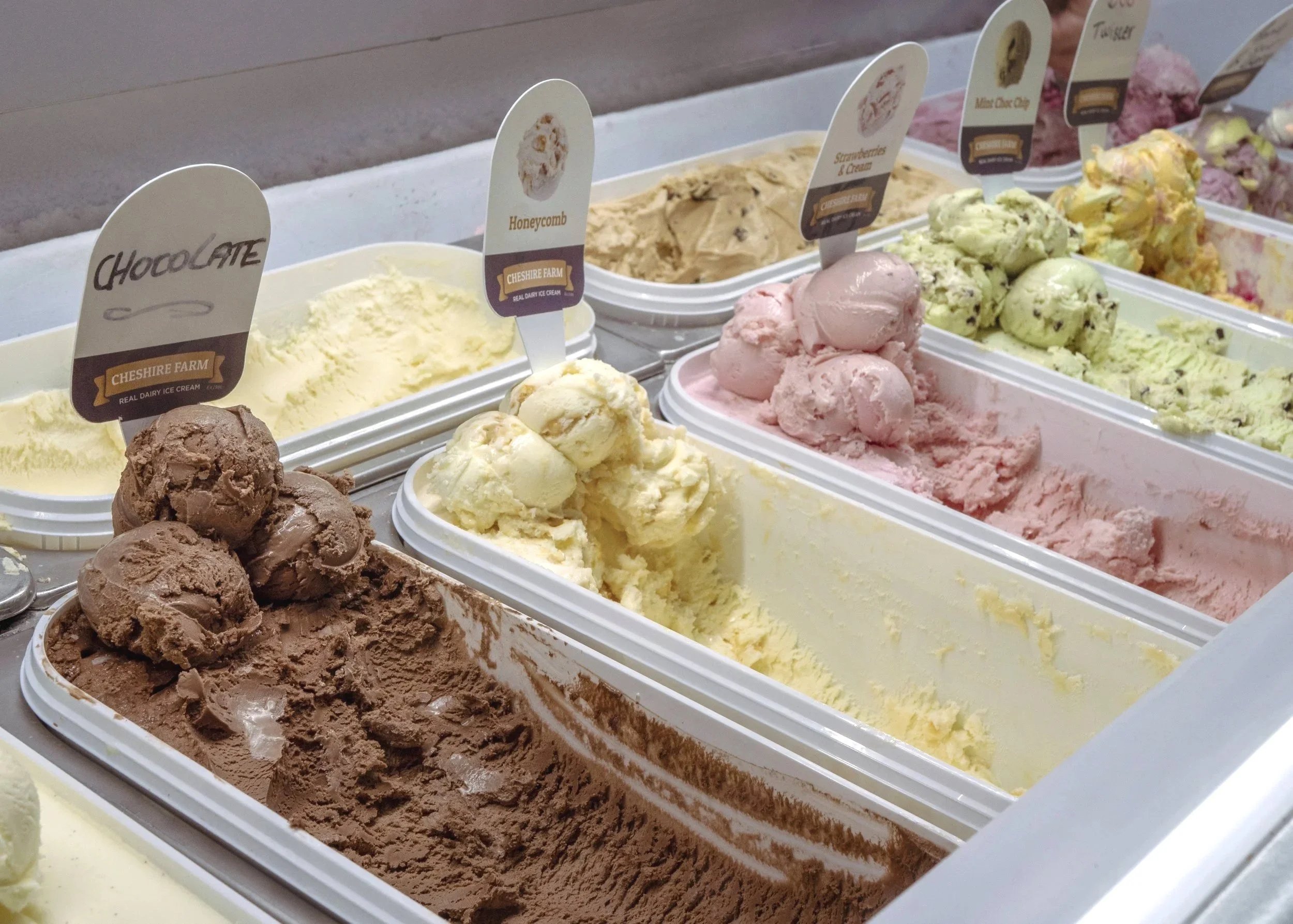 Several tubs of flavored ice cream at an ice cream shop, with flavor labels including chocolate, honeycomb, strawberry, and others.