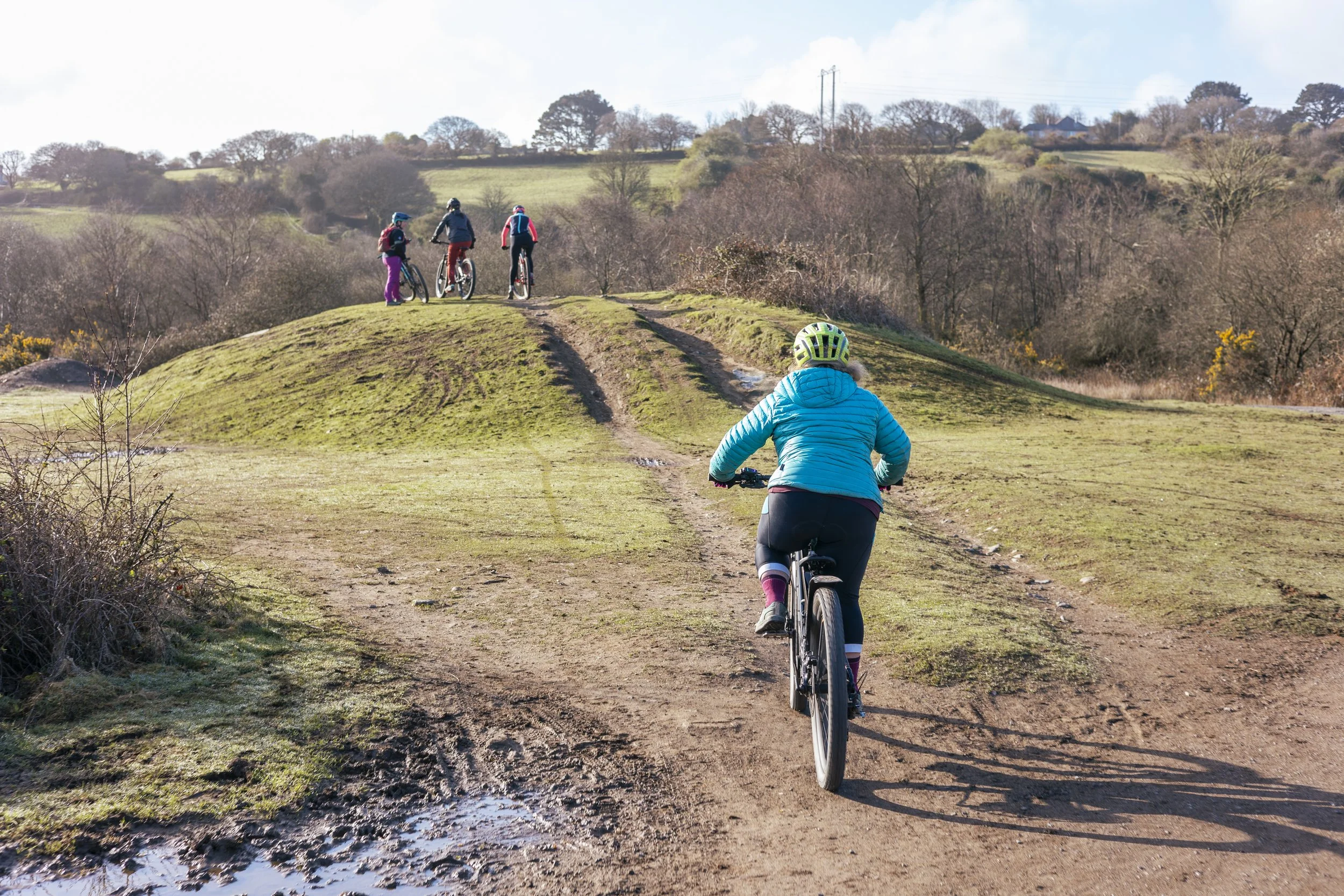A person riding a mountain bike on a dirt trail towards a group of four people with bikes standing on top of a small hill in a rural landscape with trees and open fields.