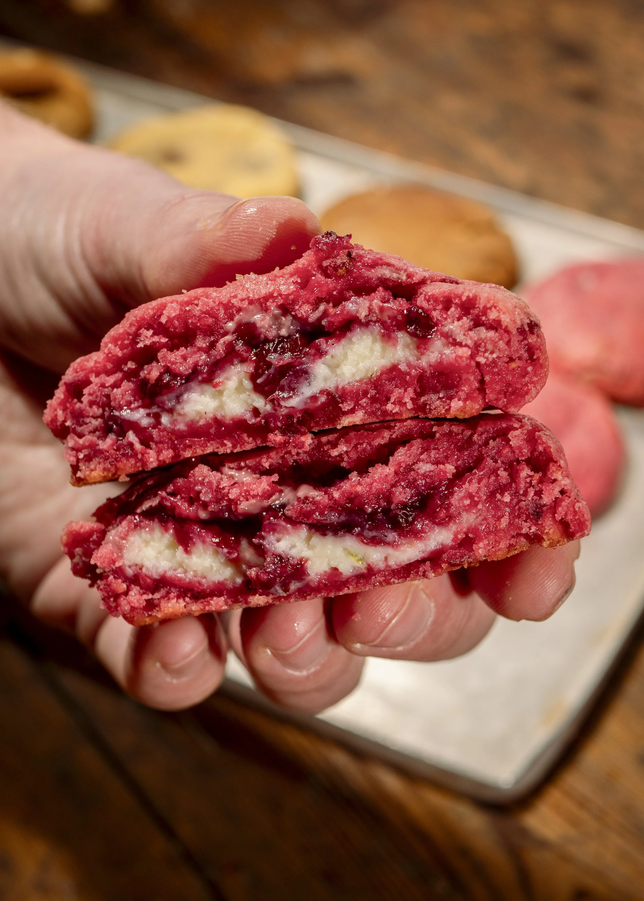 Close-up of a person holding a halved raspberry-flavored cookie filled with cream and raspberry jam, with more cookies in the background on a tray.