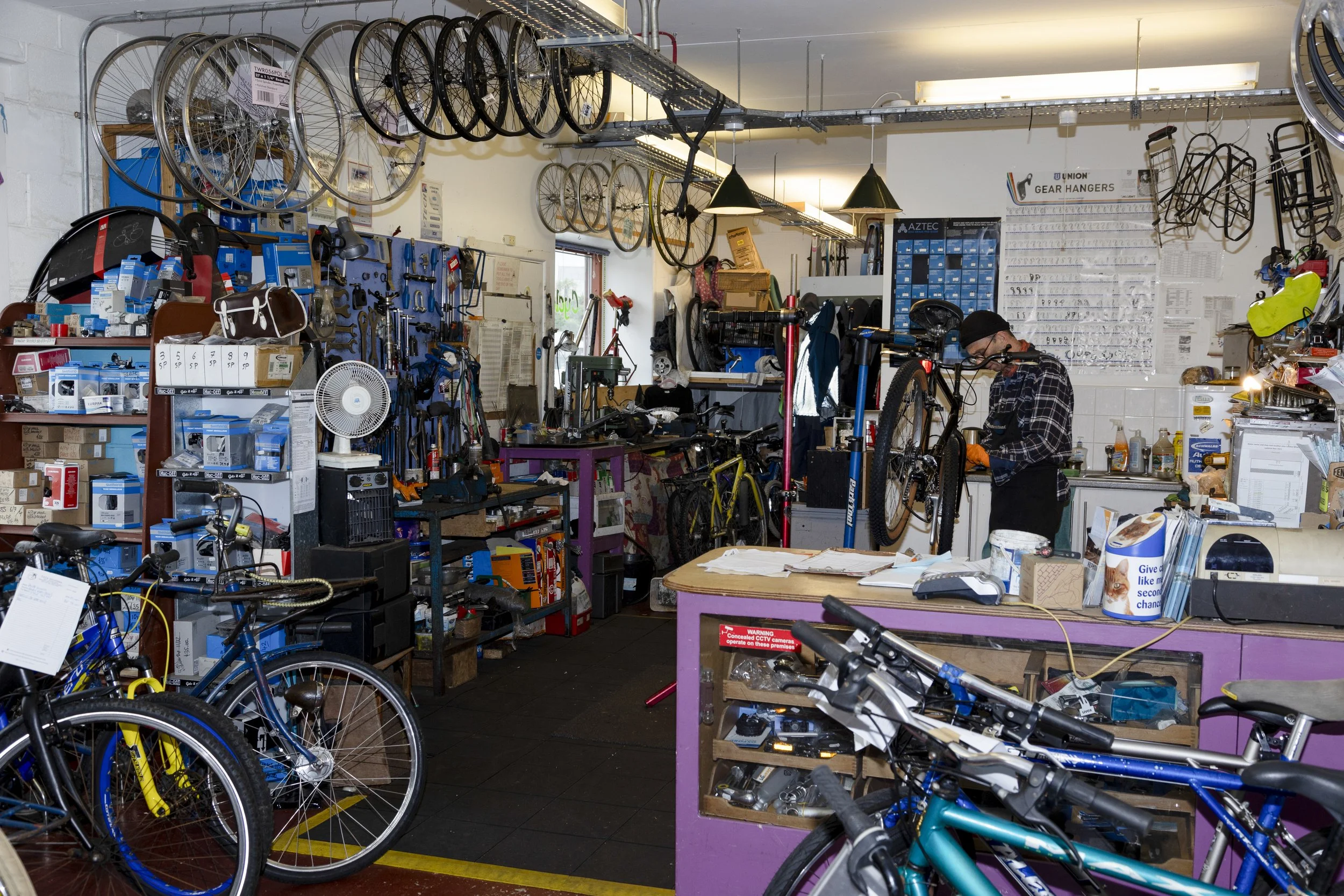 Bike shop with rows of bicycles on display, bicycle parts and accessories organized on shelves, tools hanging on walls, and a person working on a bicycle inside the store.
